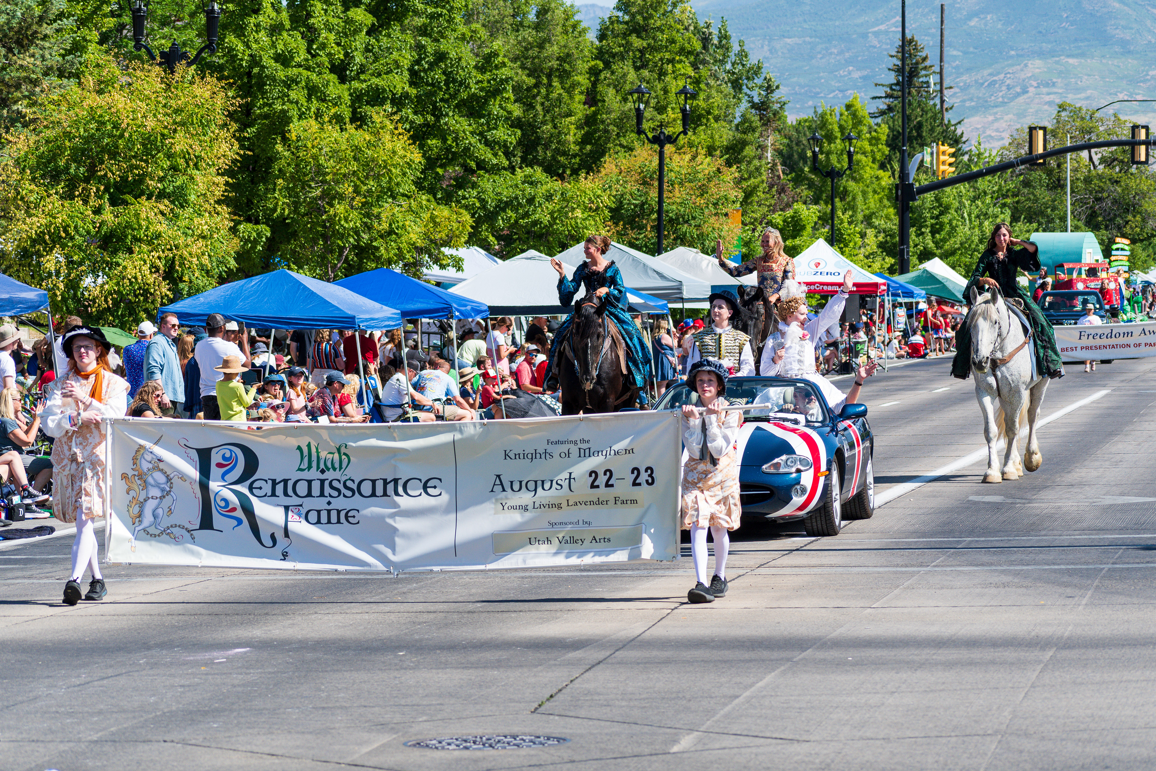 Provo, Utah – July 4, 2025: Two girls in Elizabethan costumes carry a “Utah Renaissance Faire” banner ahead of a classic Austin Healey and three women in period dress riding horses during the parade.