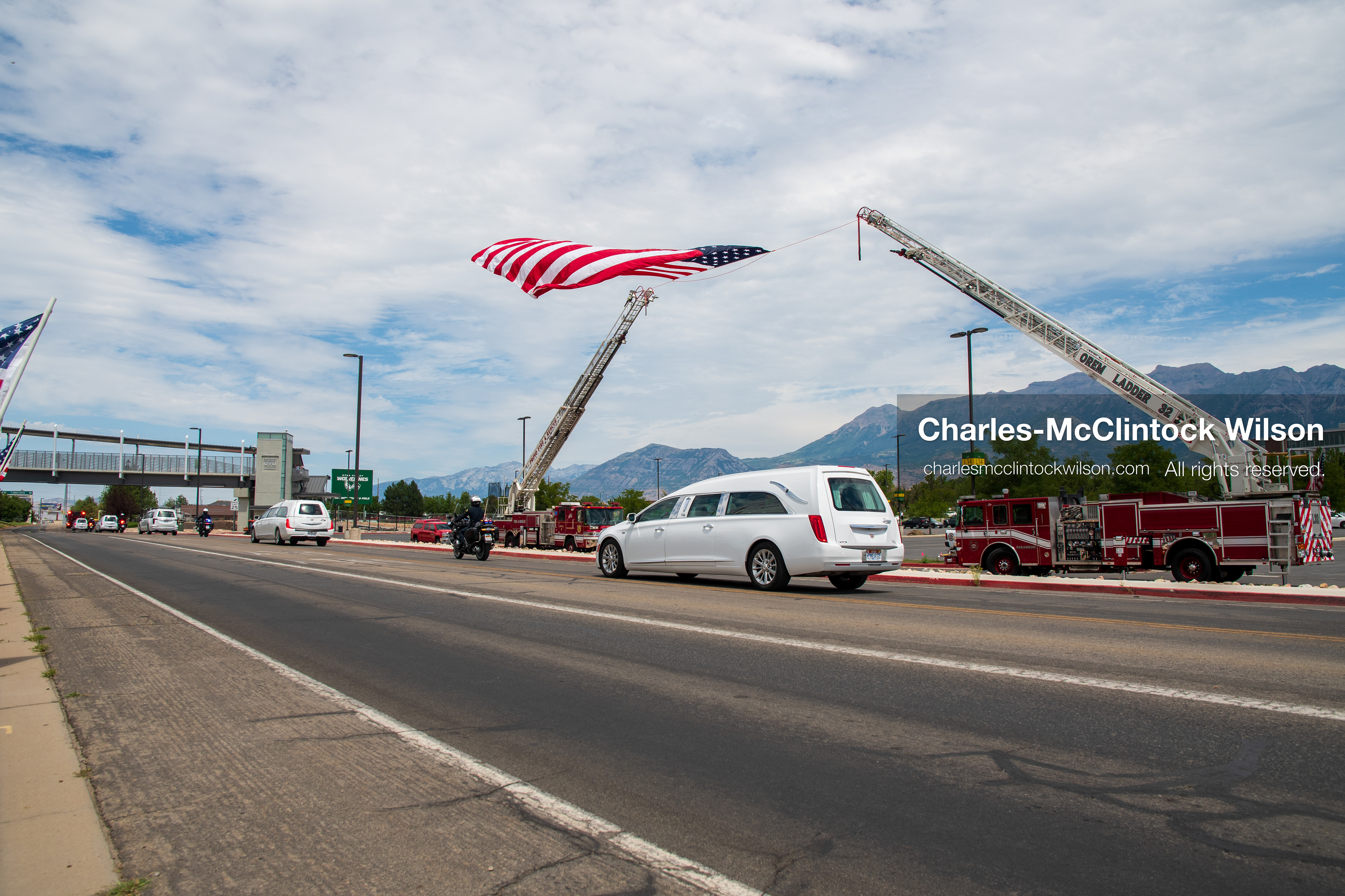 A white Cadillac hearse drives past a massive American flag suspended between two extended fire department ladder trucks during a funeral procession for Provo Fire Captain Spencer Long and his family in Orem, Utah, on Wednesday, July 29, 2026. More than 300 people came to the public memorial ceremony to honor the five family members who died after a flash flood hit their campsite near the Sunglow Campground in Wayne County. (Photo by Charles-McClintock Wilson/Sipa USA)