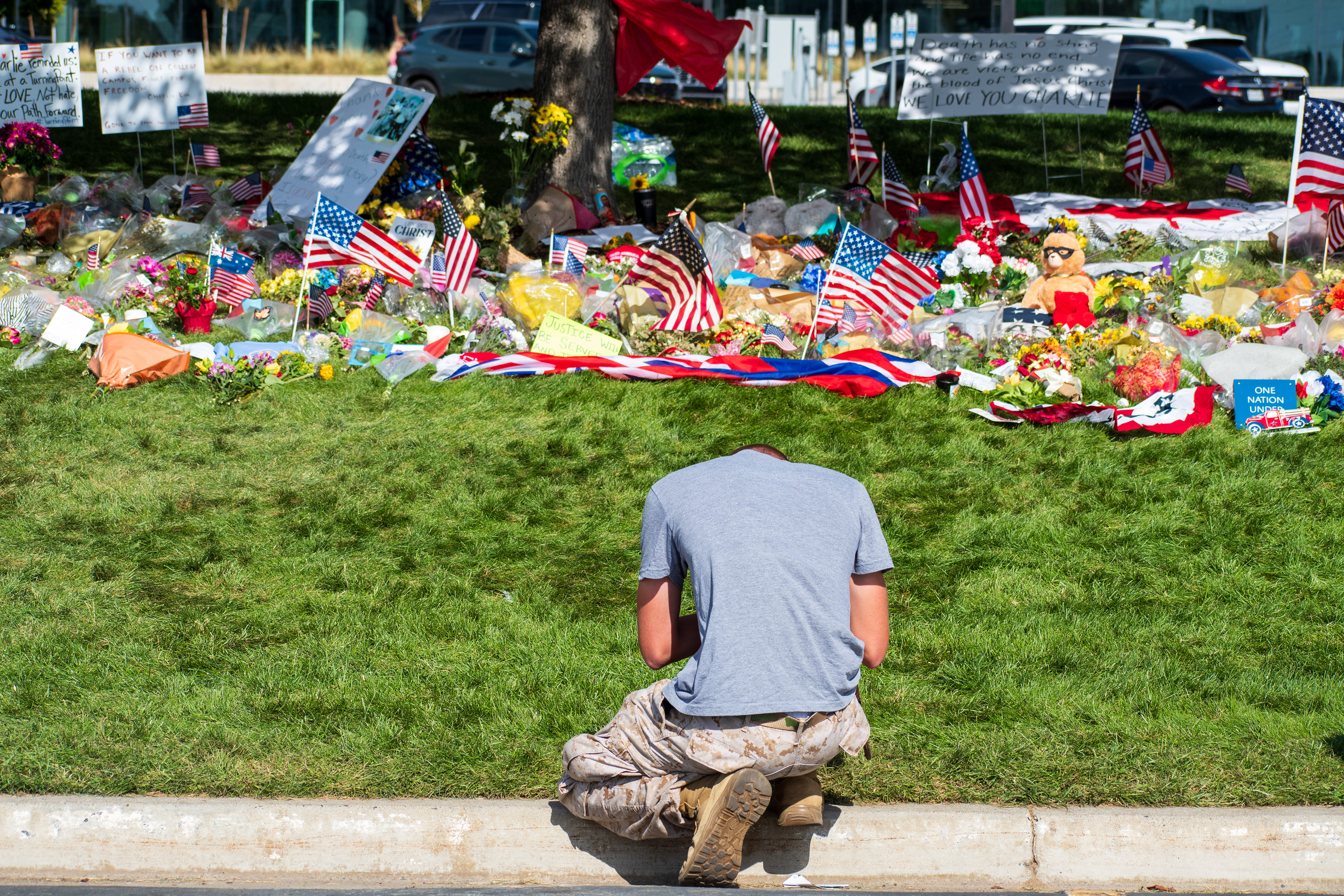 OREM, UTAH – SEPTEMBER 15, 2025: A person kneels at a memorial site for Charlie Kirk on the campus of Utah Valley University. The tribute includes American flags, flowers, stuffed animals, and handwritten signs arranged around a tree near a sidewalk. Vehicles and other individuals are visible in the background. © Charles‑McClintock Wilson / ZUMA Press