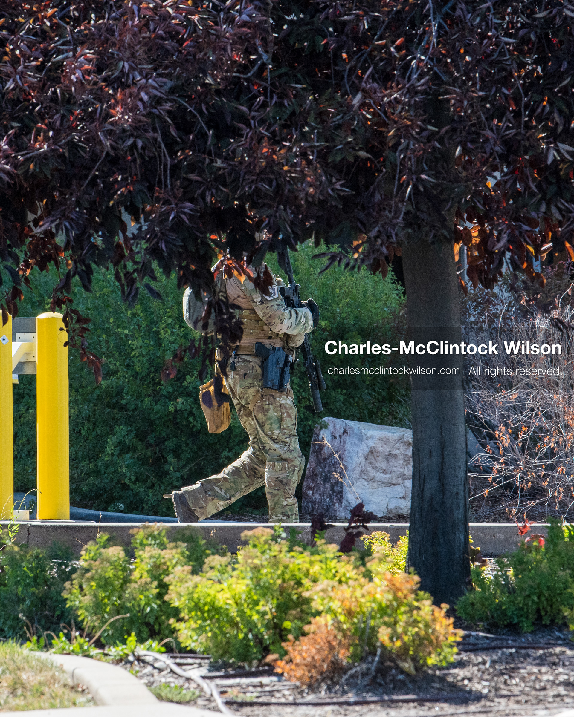 An armed law enforcement officer in camouflage tactical gear patrols a secured perimeter walkway behind foliage outside Intermountain Medical Center in Murray, Utah, on Monday, August 17, 2026. Multiple regional law enforcement agencies converged to heavily secure the hospital perimeter as a safety precaution after shooting victims arrived from a fatal shootout that erupted outside a funeral service in nearby Kearns earlier that day. (Photo by Charles-McClintock Wilson/Sipa USA)