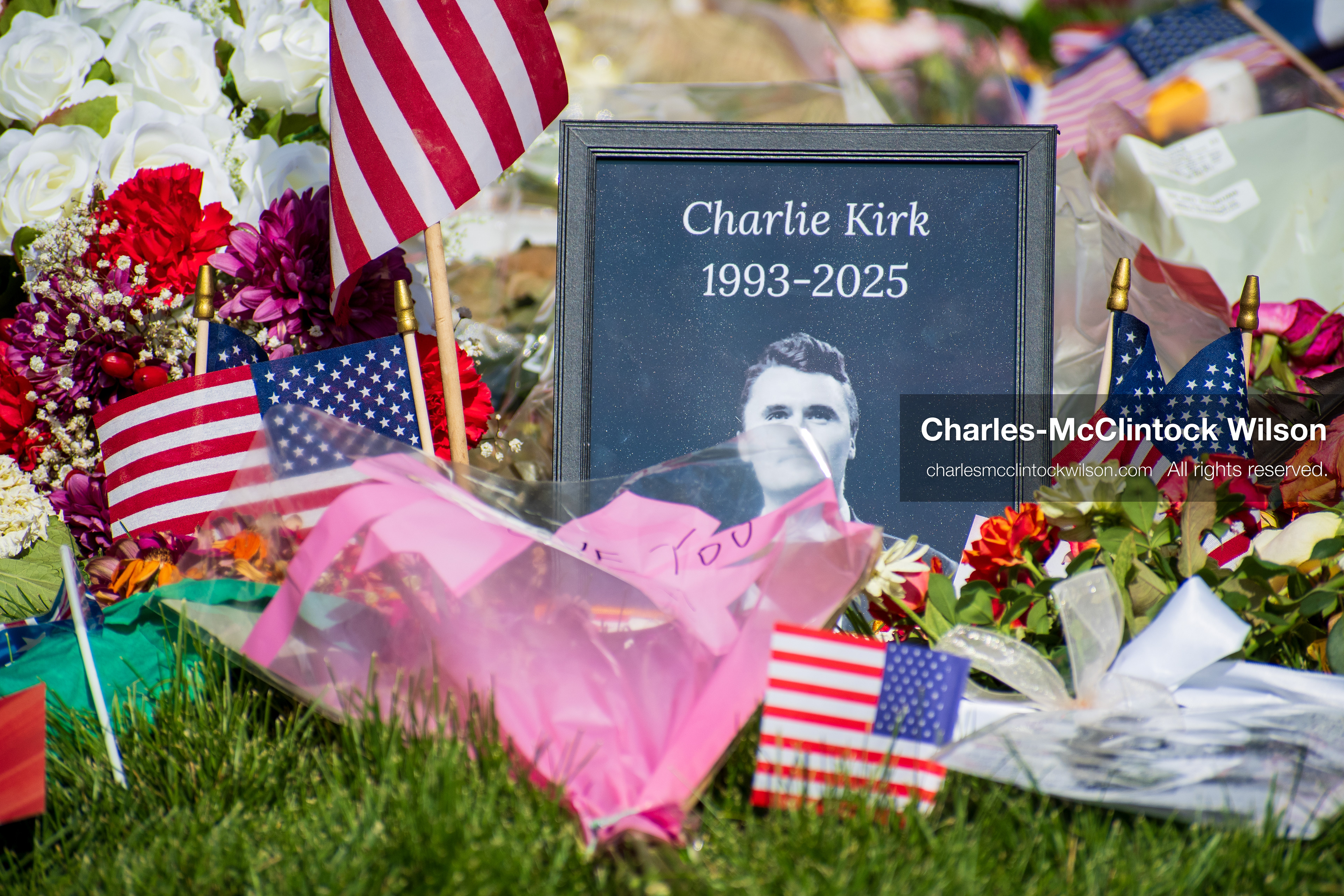 OREM, UTAH – SEPTEMBER 15, 2025: A framed photograph labeled “Charlie Kirk 1993–2025” is displayed at a memorial site on the campus of Utah Valley University. American flags, flowers, and tribute items are arranged on the grass surrounding the portrait. © Charles‑McClintock Wilson / ZUMA Press