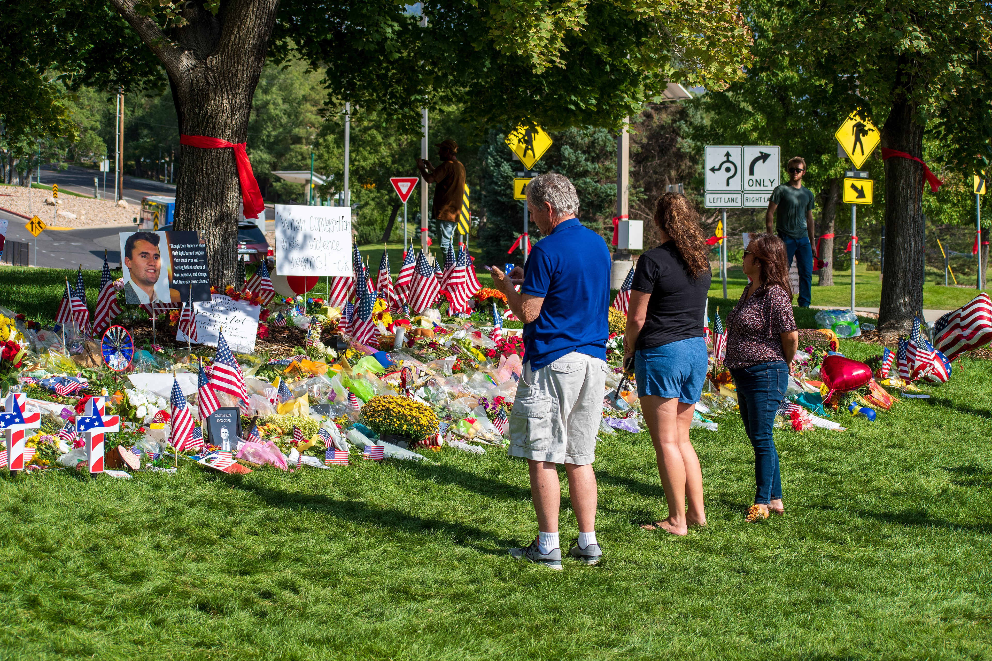 OREM, UTAH – SEPTEMBER 15, 2025: People gather at a memorial site for Charlie Kirk on the campus of Utah Valley University. The tribute includes American flags, flowers, candles, balloons, and handwritten signs arranged around a large portrait. Red ribbons are tied to nearby trees, and traffic signage is visible in the background. © Charles‑McClintock Wilson / ZUMA Press