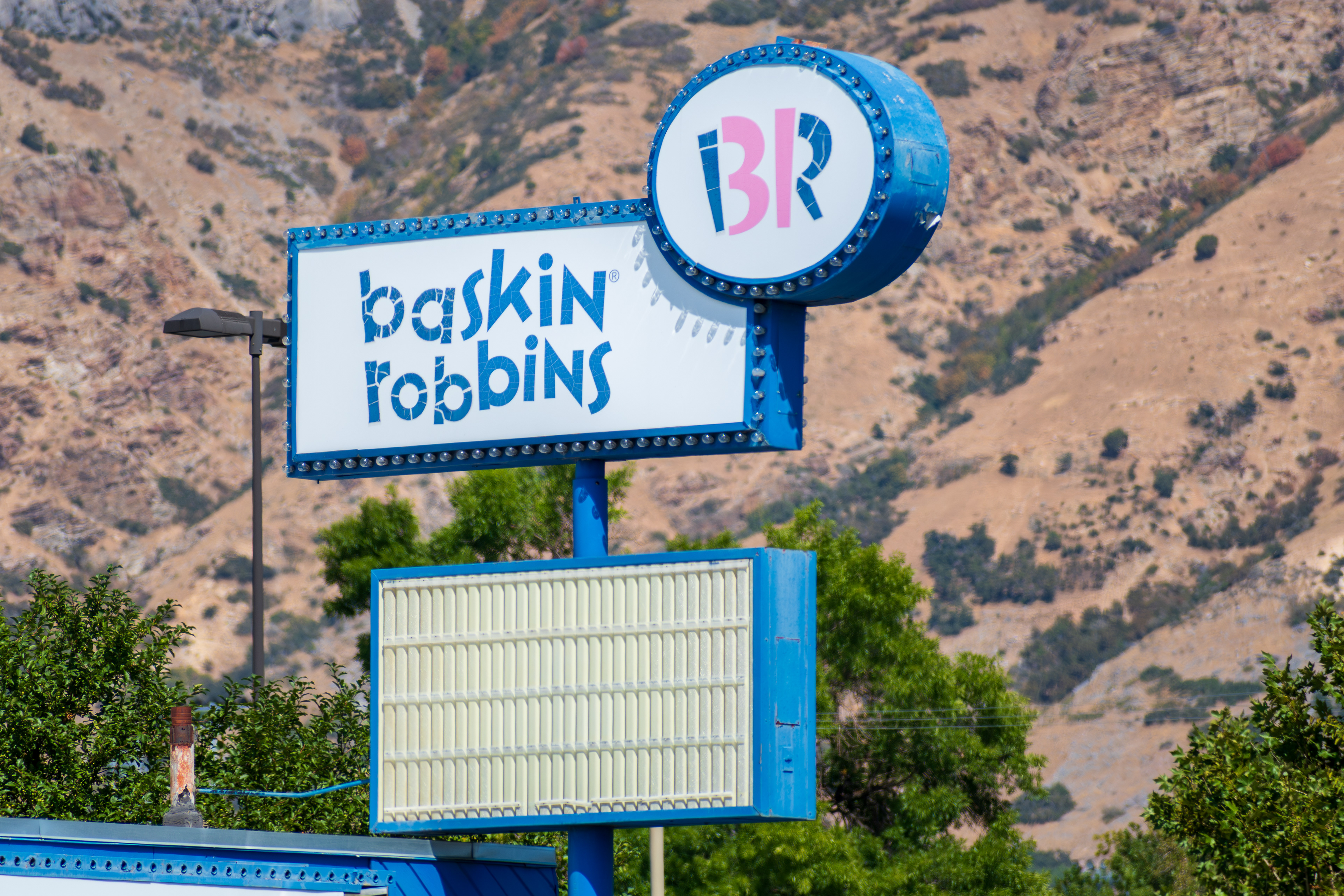 Provo, Utah, USA — September 1, 2025: A Baskin-Robbins sign stands on a blue pole with the brand’s pink and blue logo, set against a backdrop of trees and mountain ridges in central Utah. 