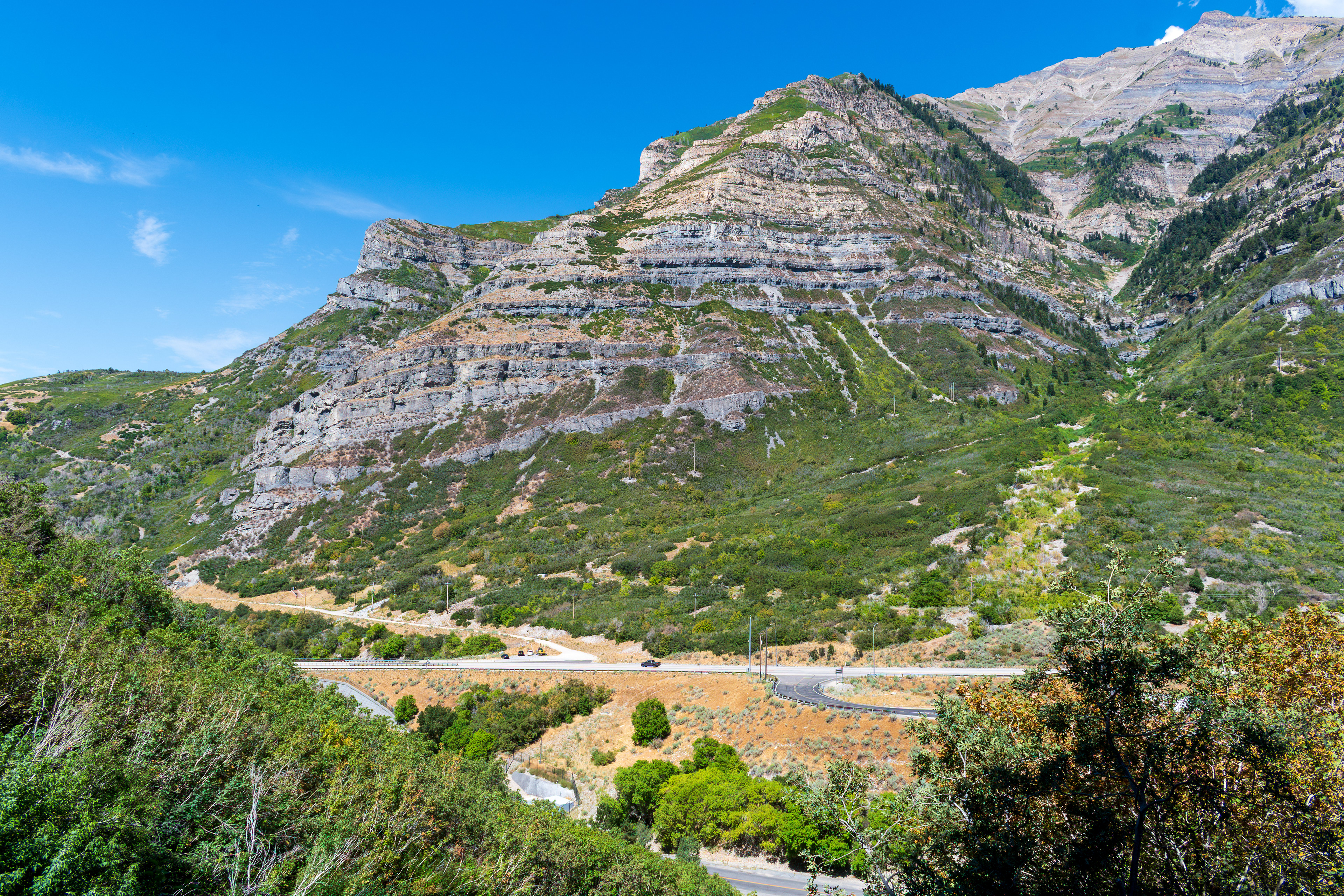 Provo, Utah, US — Sept 1, 2025: A scenic view from Bridal Veil Falls captures steep, layered rock formations and lush vegetation, with vehicles winding through the rugged terrain along U.S. Route 189.