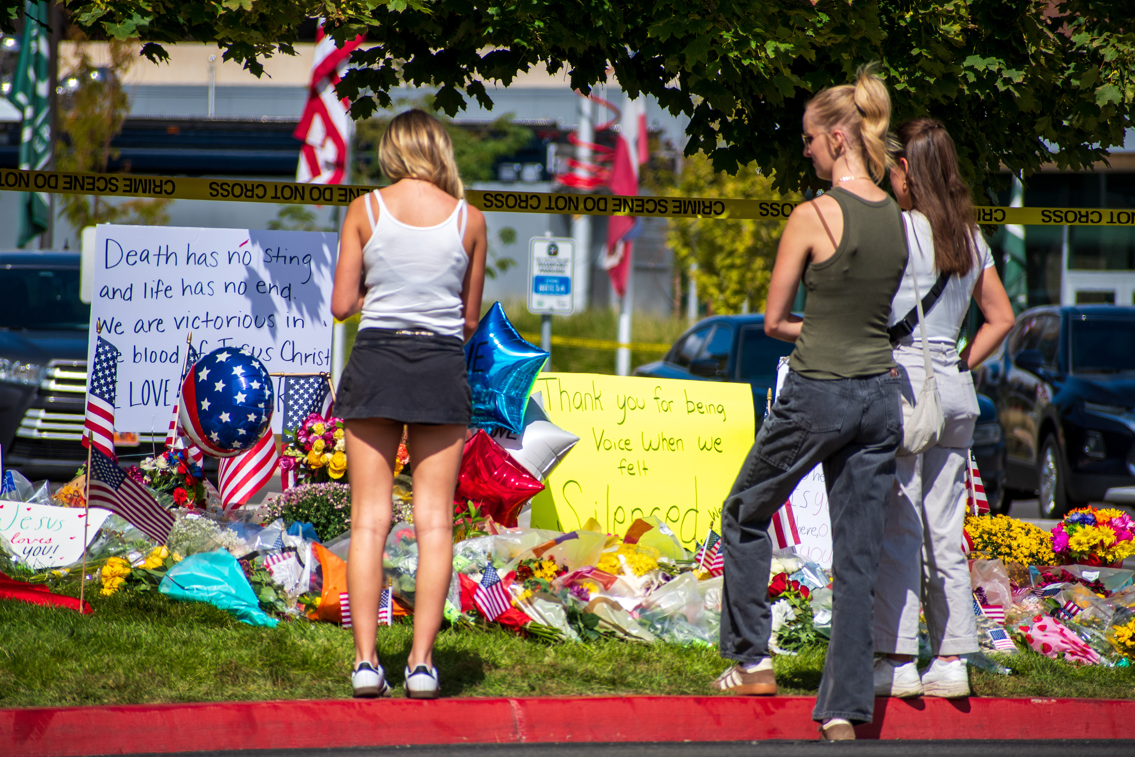 OREM, UTAH – SEPTEMBER 12, 2025: Three individuals observe a memorial site for Charlie Kirk on the campus of Utah Valley University. The tribute includes American flags, red-white-and-blue balloons, flowers, and handwritten posters. Yellow caution tape appears in the background, framing the scene with a solemn tone. © Charles‑McClintock Wilson / ZUMA Press