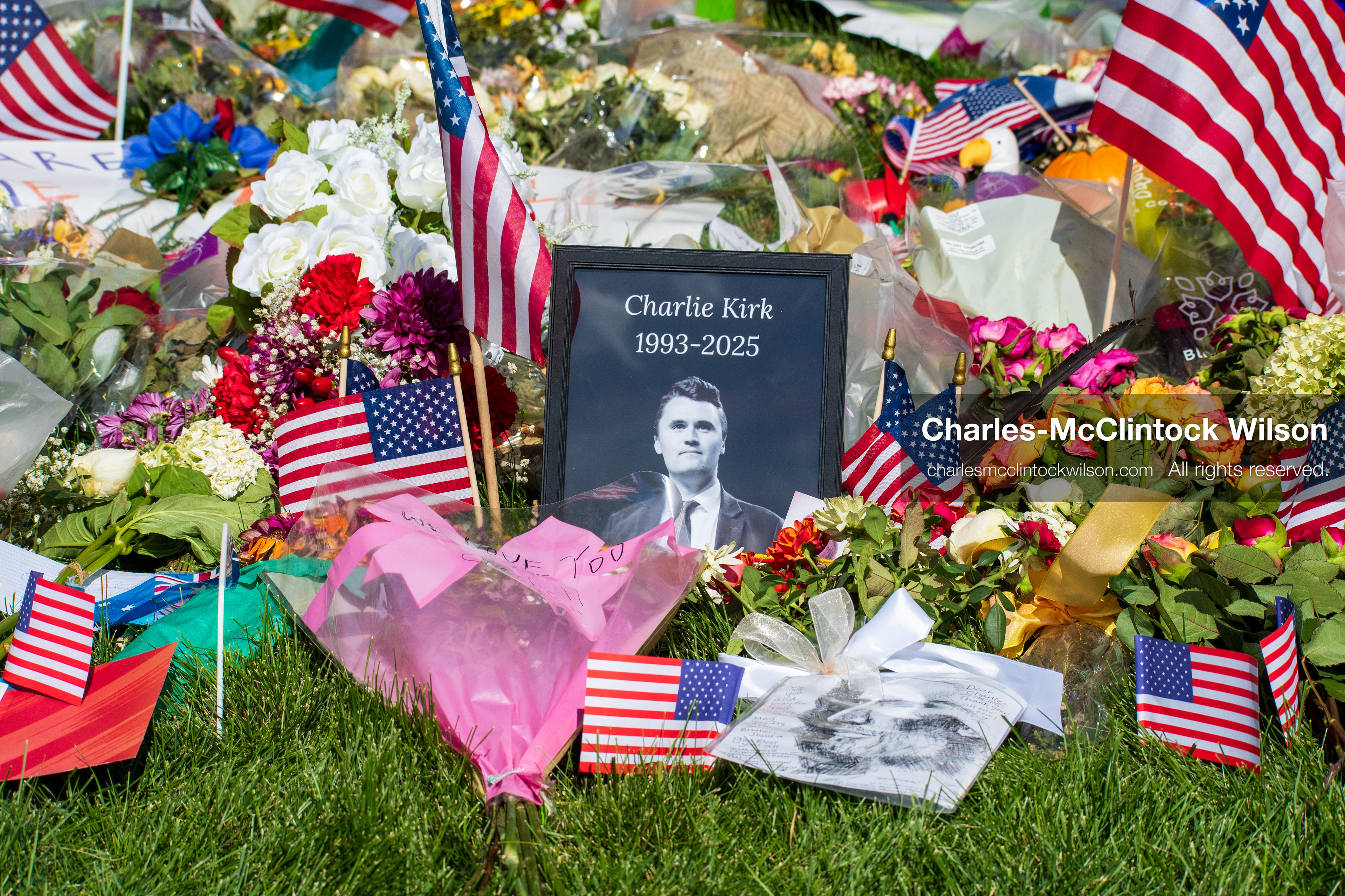 OREM, UTAH – SEPTEMBER 15, 2025: A framed photograph labeled “Charlie Kirk 1993–2025” is displayed at a memorial site on the campus of Utah Valley University. American flags, flowers, and tribute items are arranged on the grass surrounding the portrait. © Charles‑McClintock Wilson / ZUMA Press