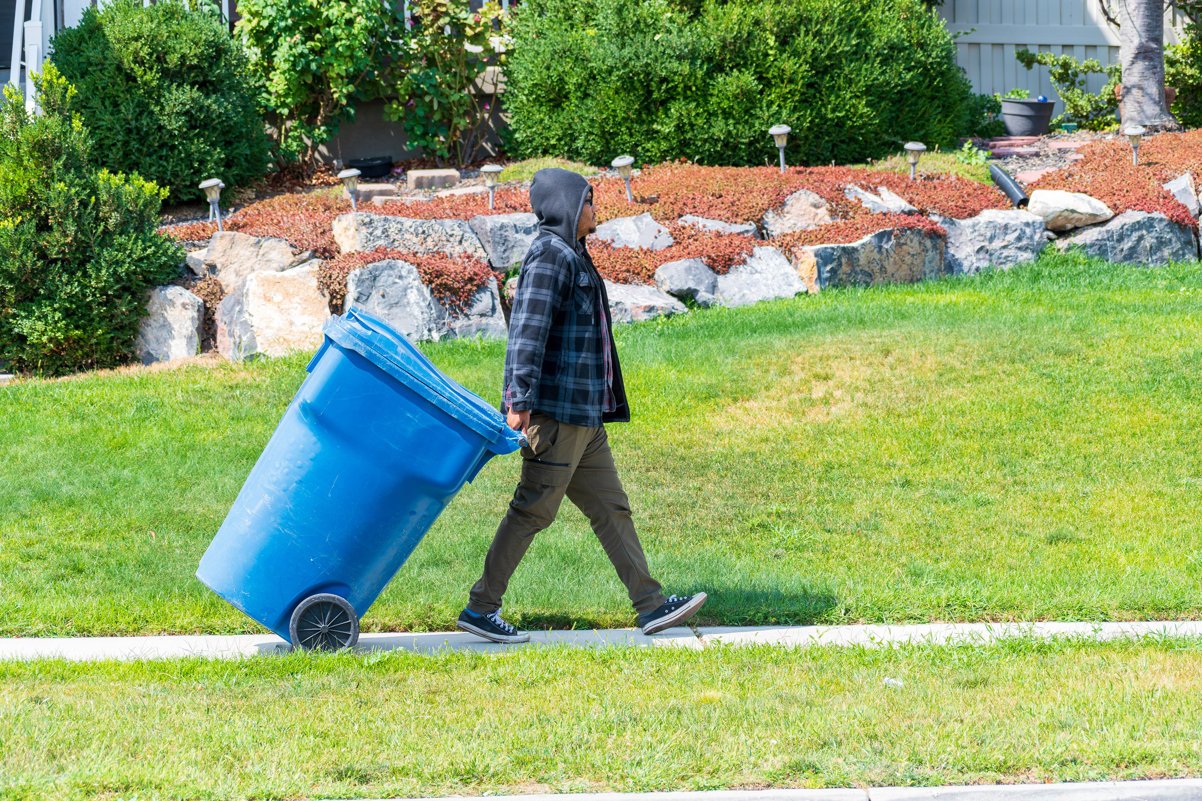 Salt Lake City, Utah, USA — September 1, 2025: A person pulls a blue recycling bin along a sidewalk bordered by grass, rocks, and a decorative metal fence.