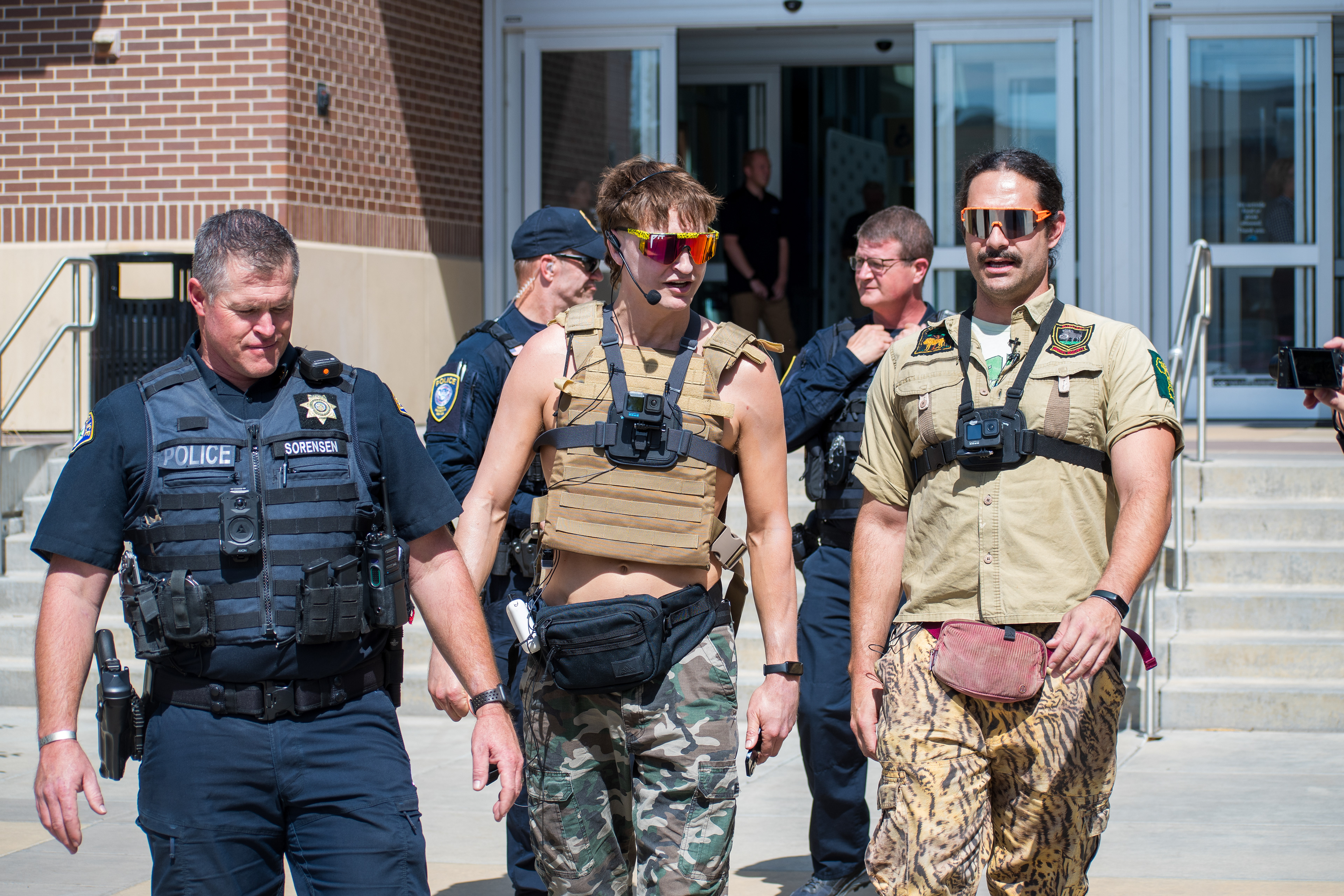 September 15, 2025 – Provo, Utah, United States: Law enforcement officers escort a young man wearing tactical gear—including a tan vest, camouflage pants, and a chest-mounted camera—outside the Utah Valley Convention Center during the Department of Homeland Security career expo. The scene reflects heightened security and the complex interplay between performative dissent and institutional response. Photograph by Charles‑McClintock Wilson / ZUMA Press Wire