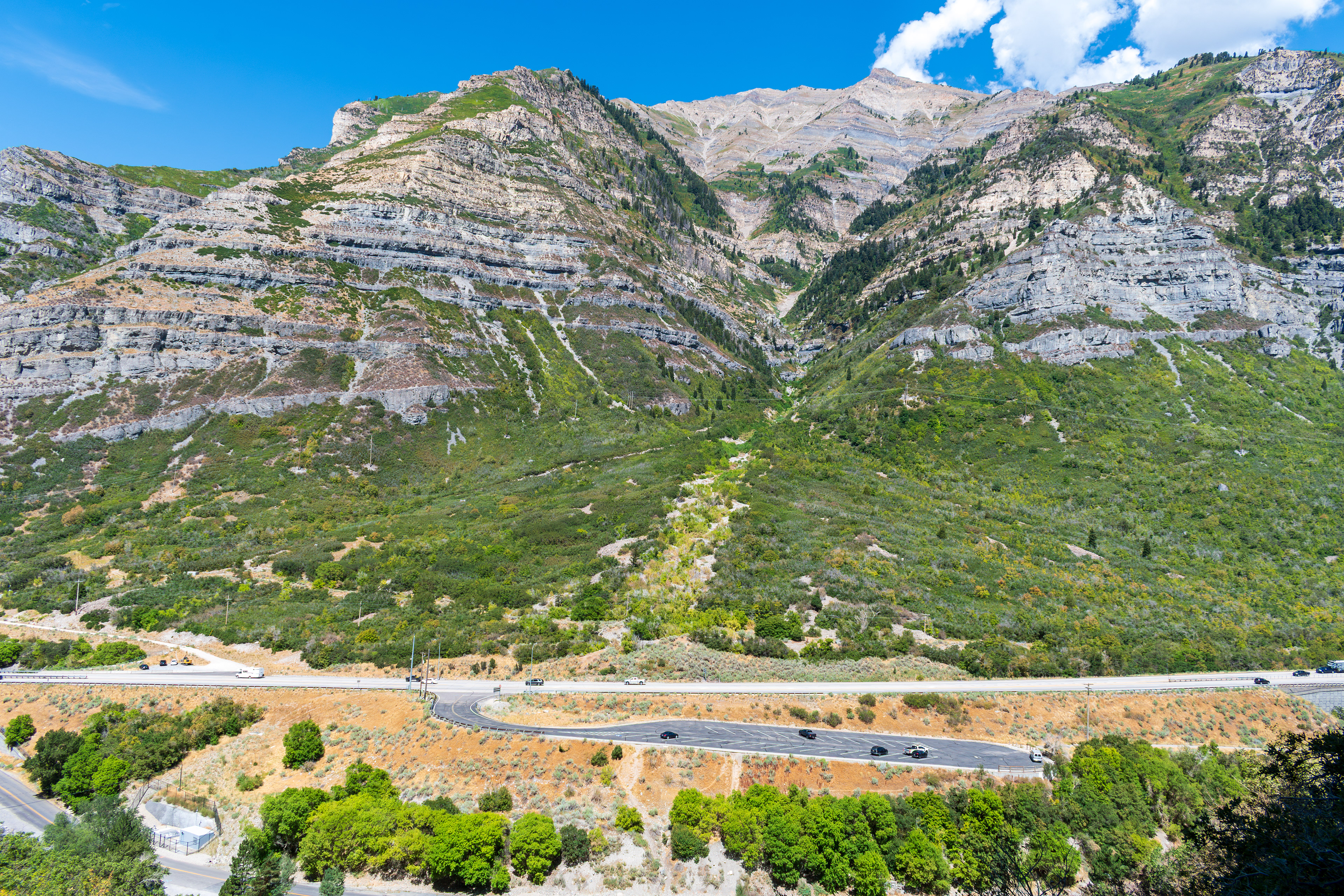 Provo, Utah, US — Sept 1, 2025: A scenic view from Bridal Veil Falls captures steep, layered rock formations and lush vegetation, with vehicles winding through the rugged terrain along U.S. Route 189.