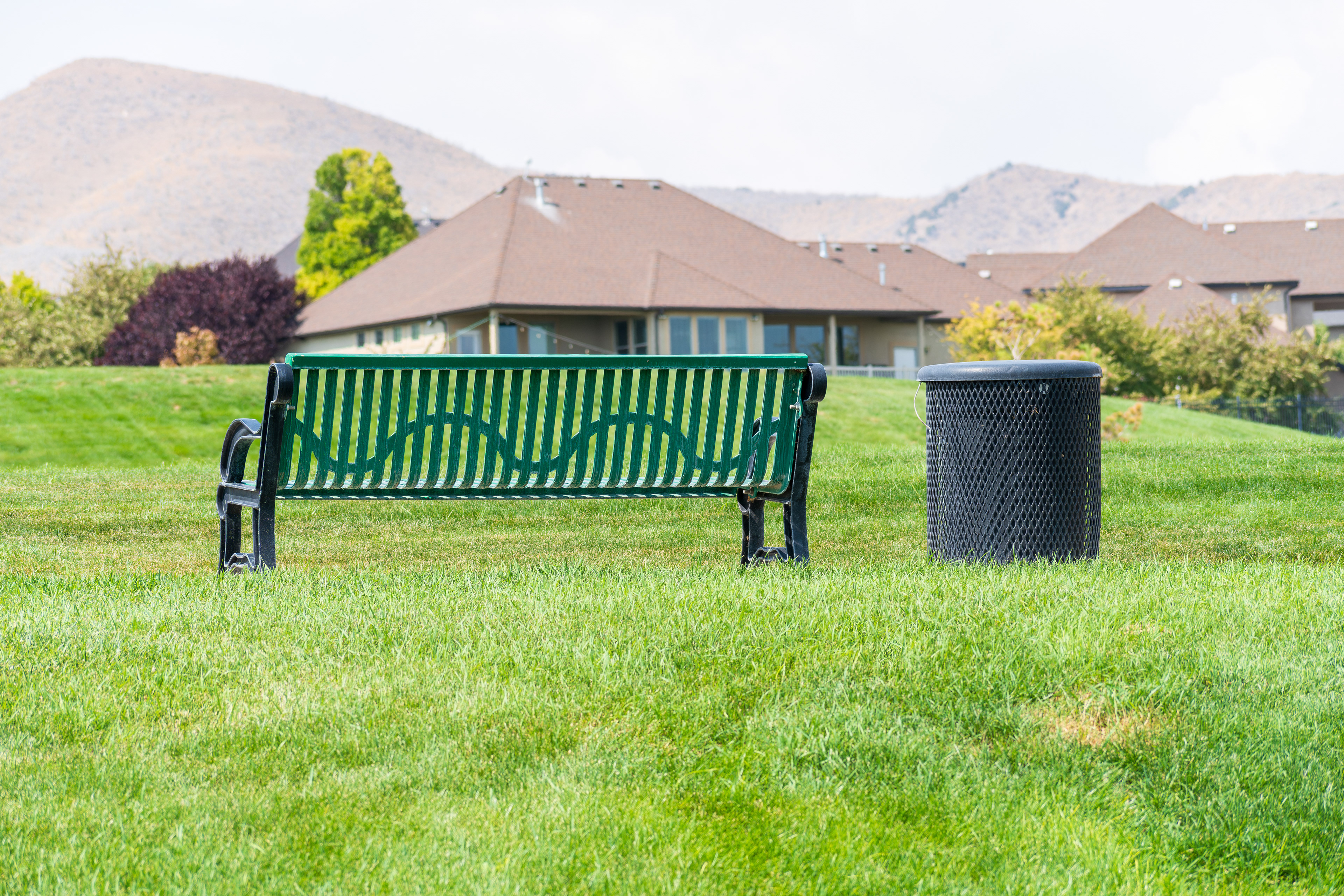 Salt Lake City, Utah, USA — September 1, 2025: A green metal park bench sits on a grassy area near a sidewalk with mountain terrain in the background.