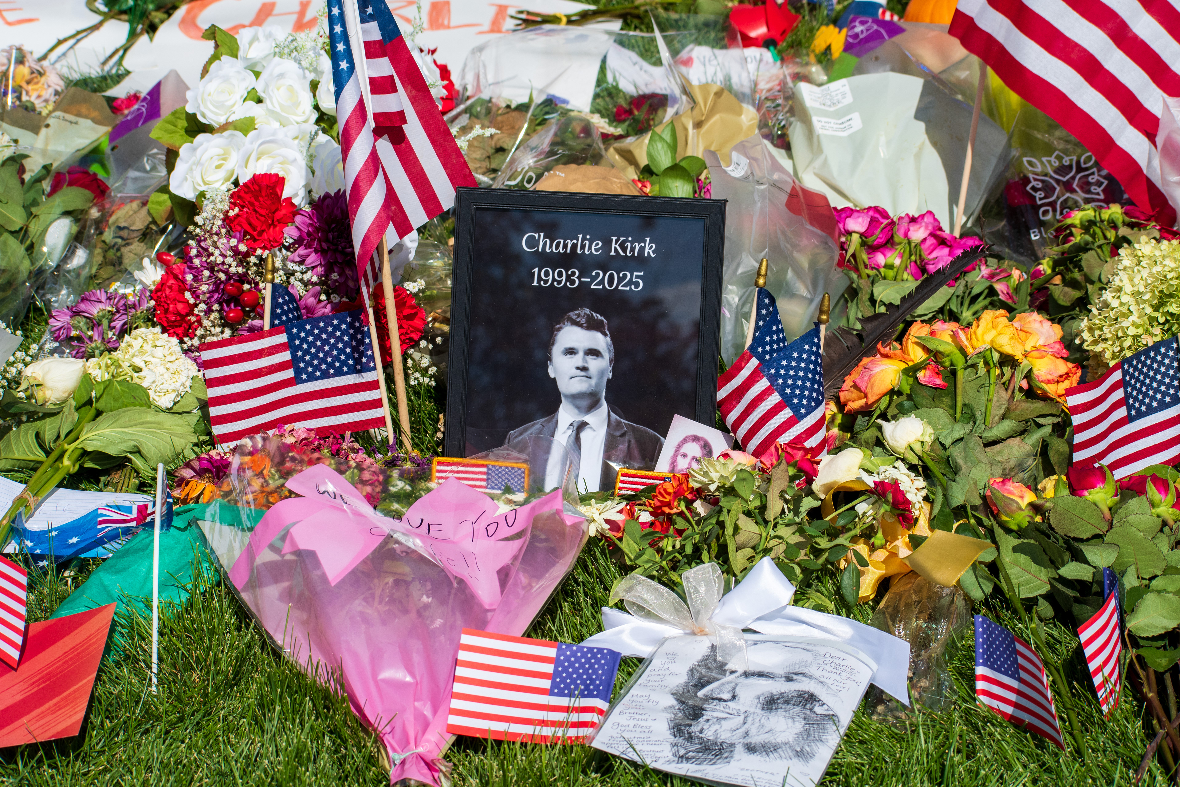 OREM, UTAH – SEPTEMBER 15, 2025: A framed photograph labeled “Charlie Kirk 1993–2025” is displayed at a memorial site on the campus of Utah Valley University. American flags, flowers, and tribute items are arranged on the grass surrounding the portrait. © Charles‑McClintock Wilson / ZUMA Press