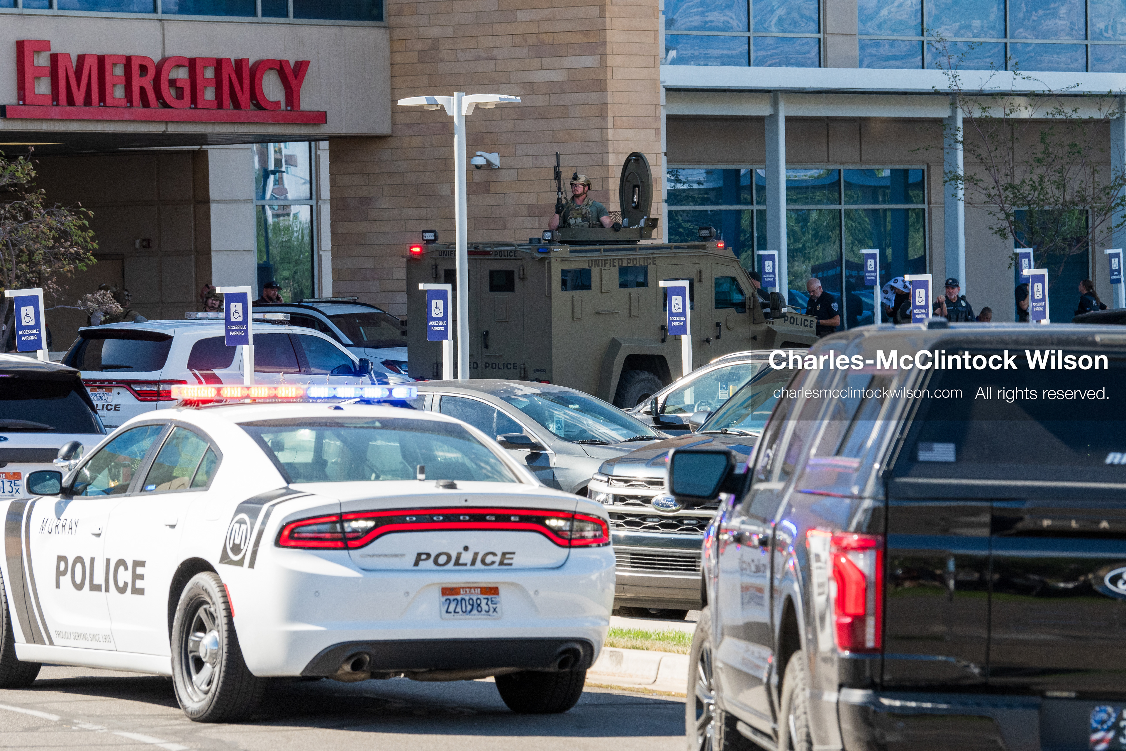 An armed law enforcement officer stands inside the roof turret hatch of an armored tactical vehicle positioned near the emergency department entrance at Intermountain Medical Center in Murray, Utah, United States, on August 17, 2026. Multiple regional police agencies secured the hospital perimeter as a safety precaution after shooting victims were admitted following a fatal shootout outside a funeral service in nearby Kearns. (Photo by Charles-McClintock Wilson/NurPhoto)