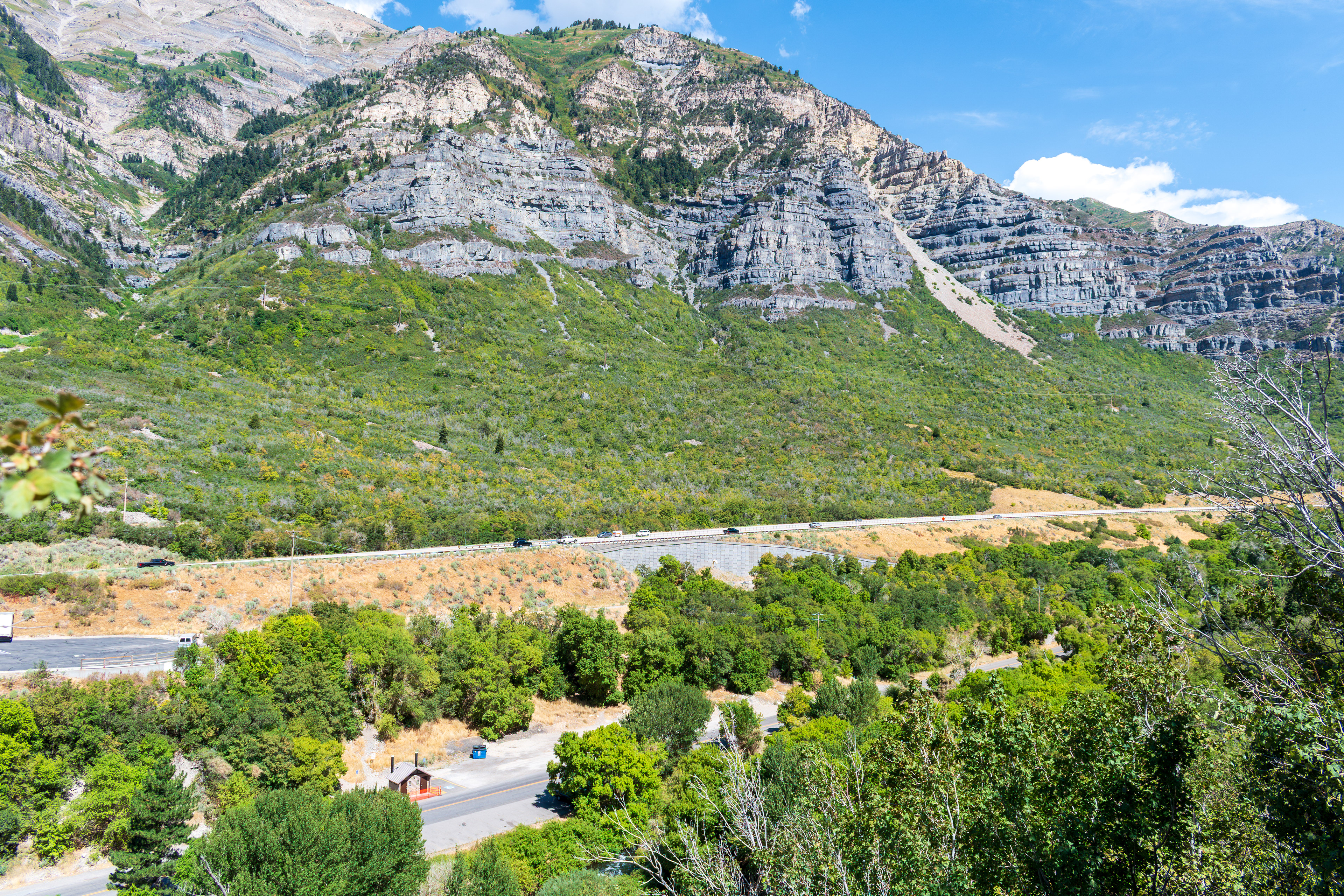 Provo, Utah, US — Sept 1, 2025: A scenic view from Bridal Veil Falls captures steep, layered rock formations and lush vegetation, with vehicles winding through the rugged terrain along U.S. Route 189.