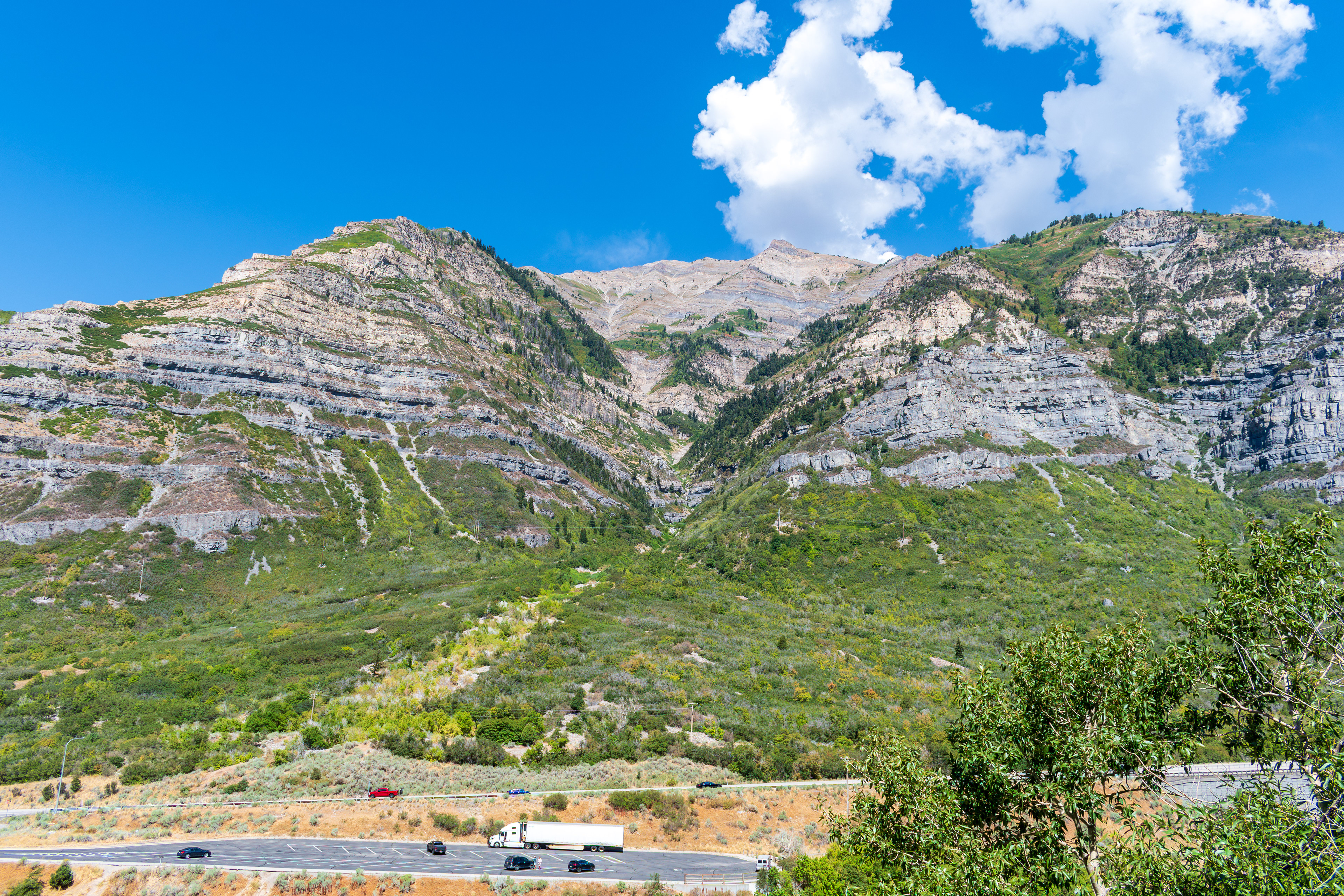 Provo, Utah, US — Sept 1, 2025: A scenic view from Bridal Veil Falls captures steep, layered rock formations and lush vegetation, with vehicles winding through the rugged terrain along U.S. Route 189.