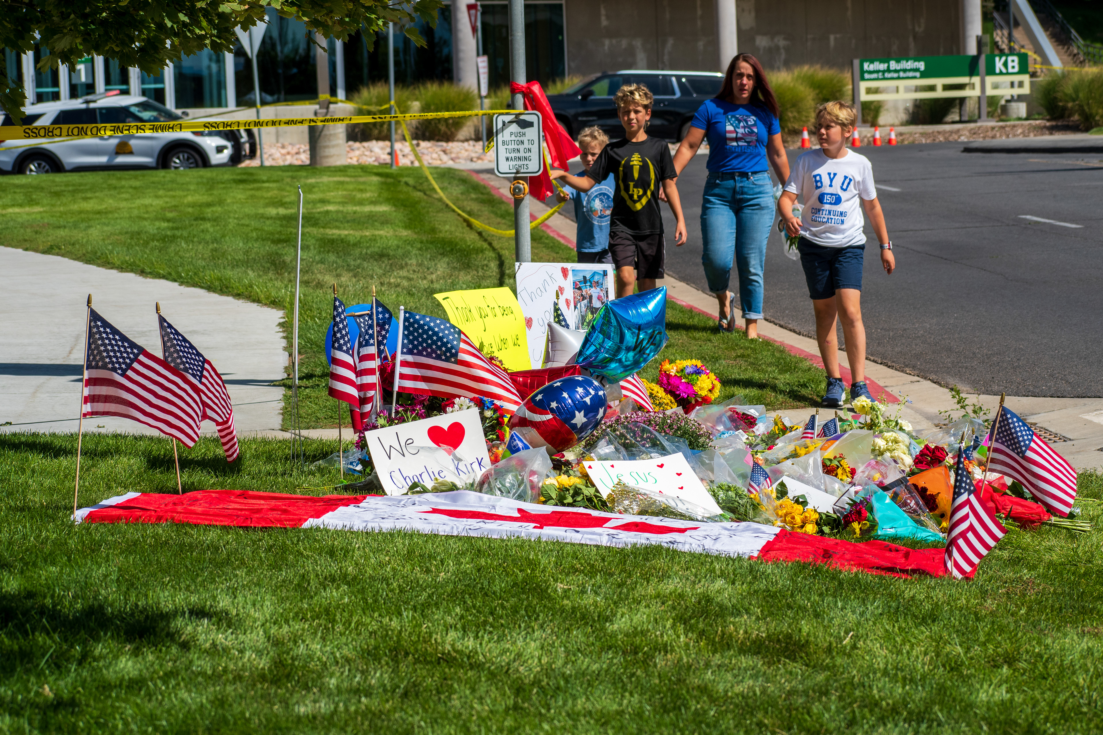 OREM, UTAH – SEPTEMBER 12, 2025: A memorial site for Charlie Kirk is seen on the lawn beside the Keller Building at Utah Valley University. American flags, flowers, balloons, and handwritten posters surround a red-and-white cloth laid out among the tributes. Two adults and two children walk toward the display. © Charles‑McClintock Wilson / ZUMA Press