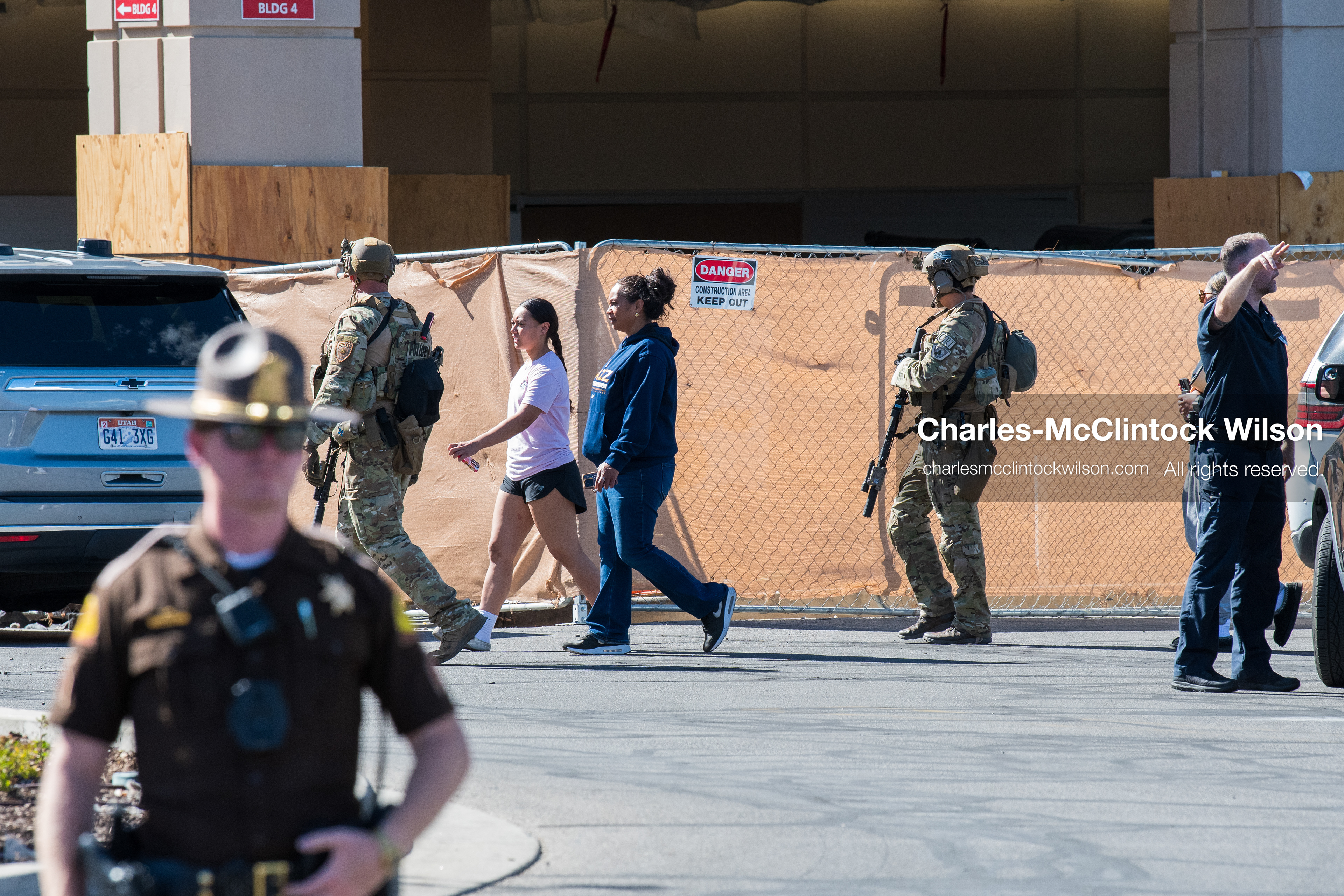 A Utah Highway Patrol trooper stands guard as individuals walk past a temporary construction barrier outside Intermountain Medical Center in Murray, Utah, United States, on August 17, 2026. Multiple regional police agencies secured the hospital perimeter as a safety precaution after shooting victims were admitted following a fatal shootout outside a funeral service in nearby Kearns. (Photo by Charles-McClintock Wilson/NurPhoto)