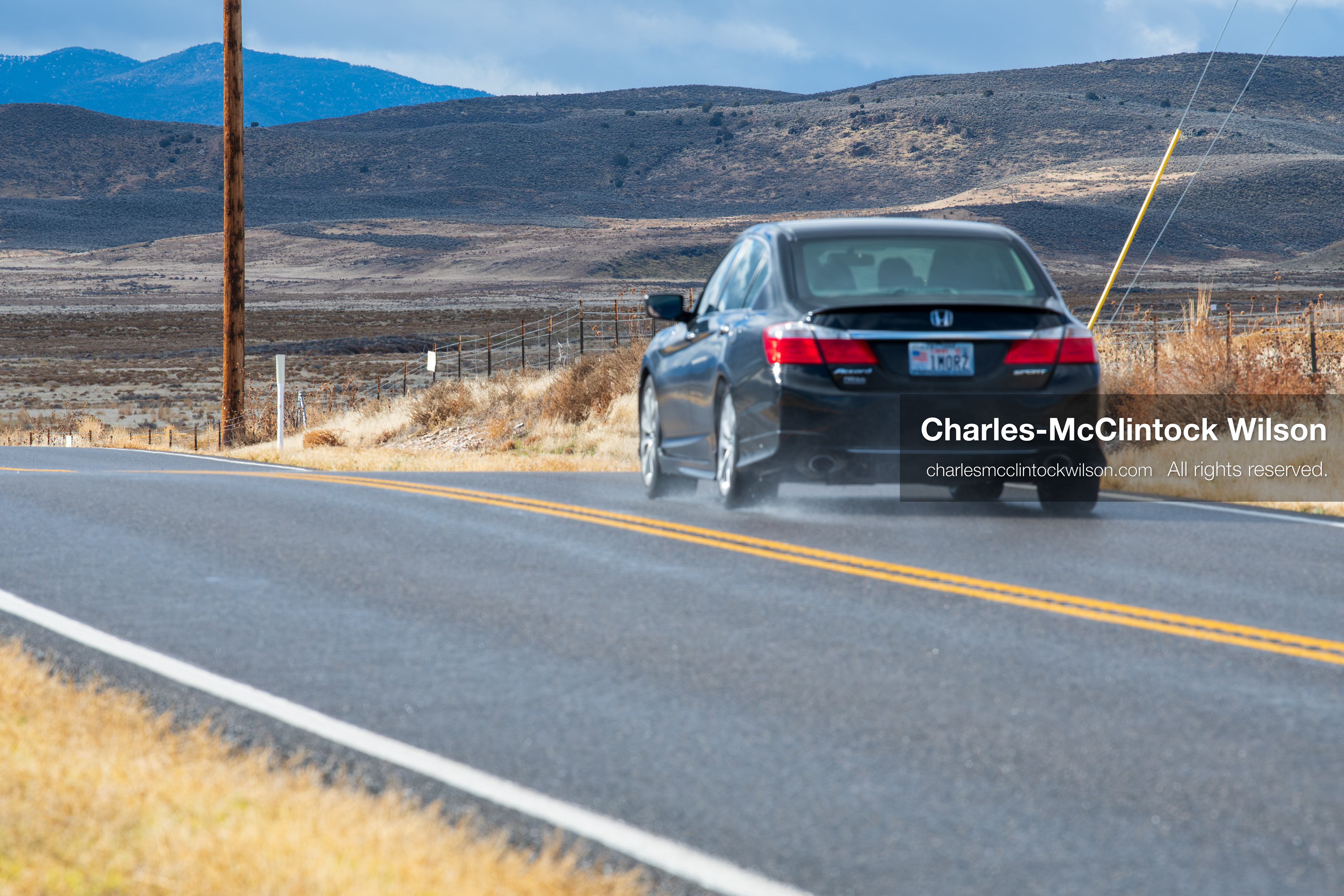 January 1, 2026, Saratoga Springs, Utah, USA: A black sedan travels along a rural road near Utah Lake on January 1, 2026, in Saratoga Springs, Utah, USA. Tire spray reflects wet conditions as dry grass, utility poles, and distant mountains frame the landscape under partly cloudy skies. (Credit Image: © Charles-McClintock Wilson/ZUMA Press Wire)
