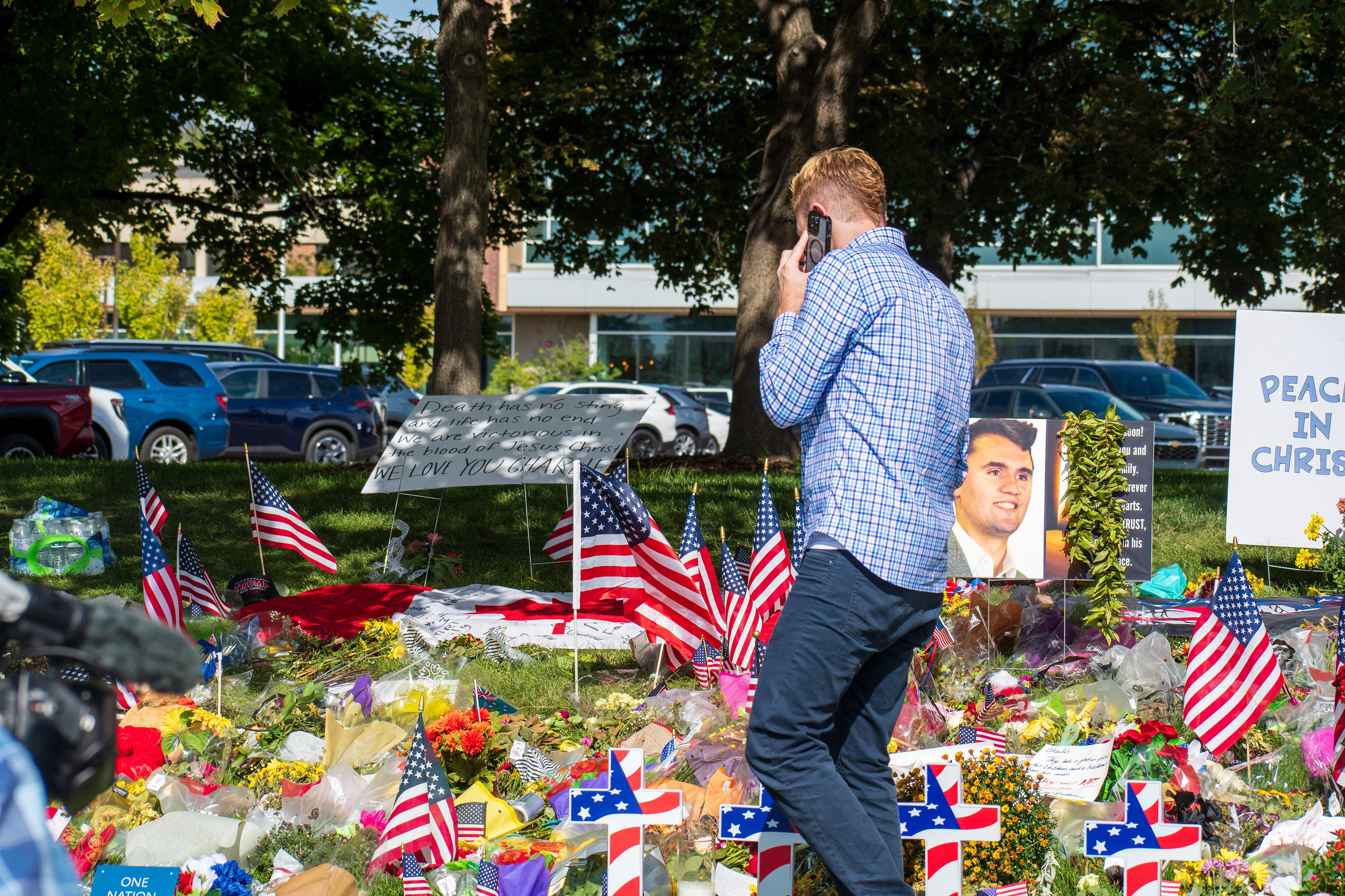 OREM, UTAH – SEPTEMBER 15, 2025: A man in a blue checkered shirt speaks on the phone while observing a memorial site for Charlie Kirk on the campus of Utah Valley University. The tribute includes American flags, flowers, candles, and a large portrait surrounded by handwritten signs. A sign reading “PEACE IN CHRIST” is visible in the background. © Charles‑McClintock Wilson / ZUMA Press