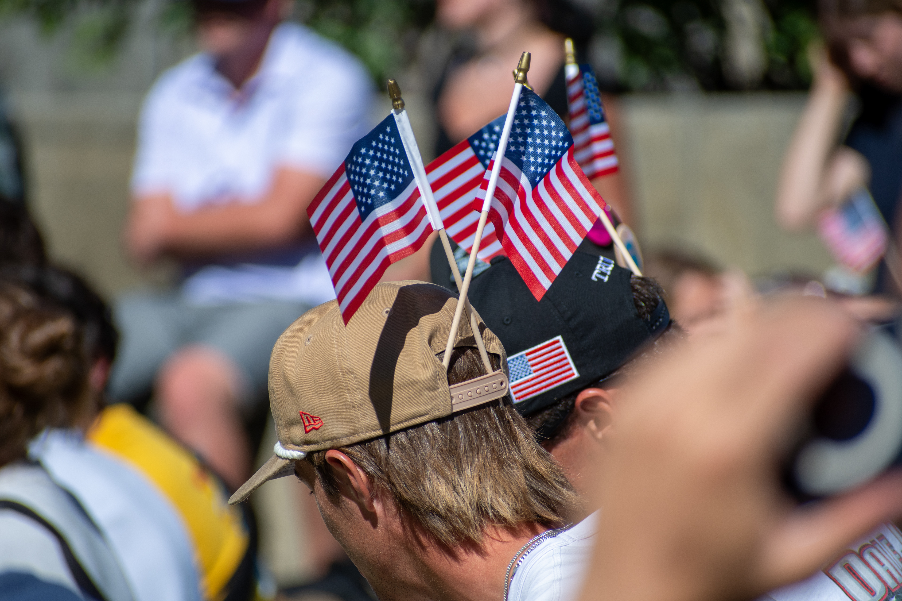 OREM, UTAH – SEPTEMBER 10, 2025: Attendees gather outdoors at Utah Valley University during the opening stop of the American Comeback Tour. Two individuals wear caps adorned with small American flags, reflecting a moment of personal expression and quiet symbolism. The image captures the warmth, texture, and communal presence that shaped the event’s atmosphere. © Charles-McClintock Wilson / ZUMA Press