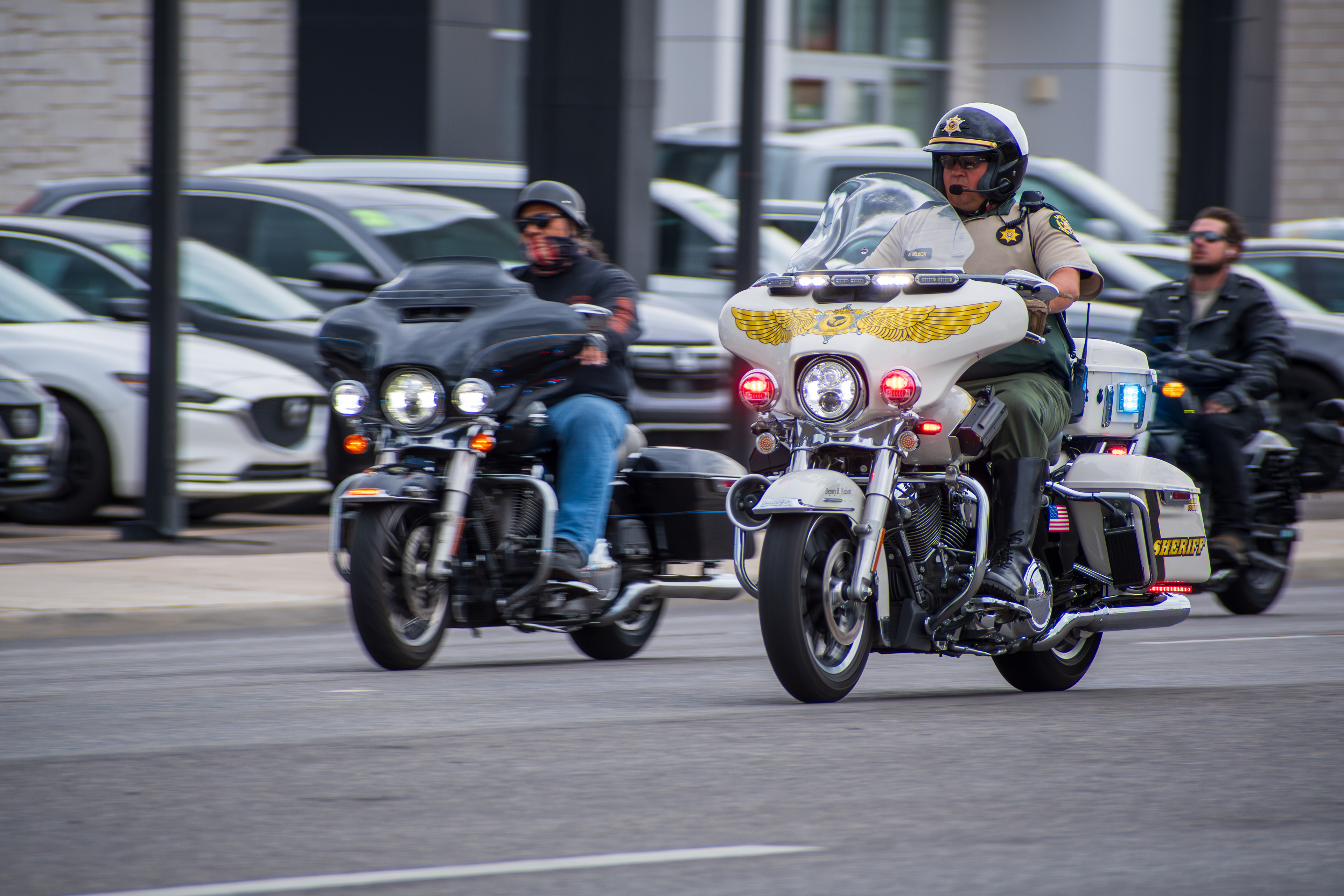 Murray, Utah — Sept. 8, 2025: A police officer and a civilian rider travel side by side on motorcycles through a city street, framed by parked cars and urban textures.