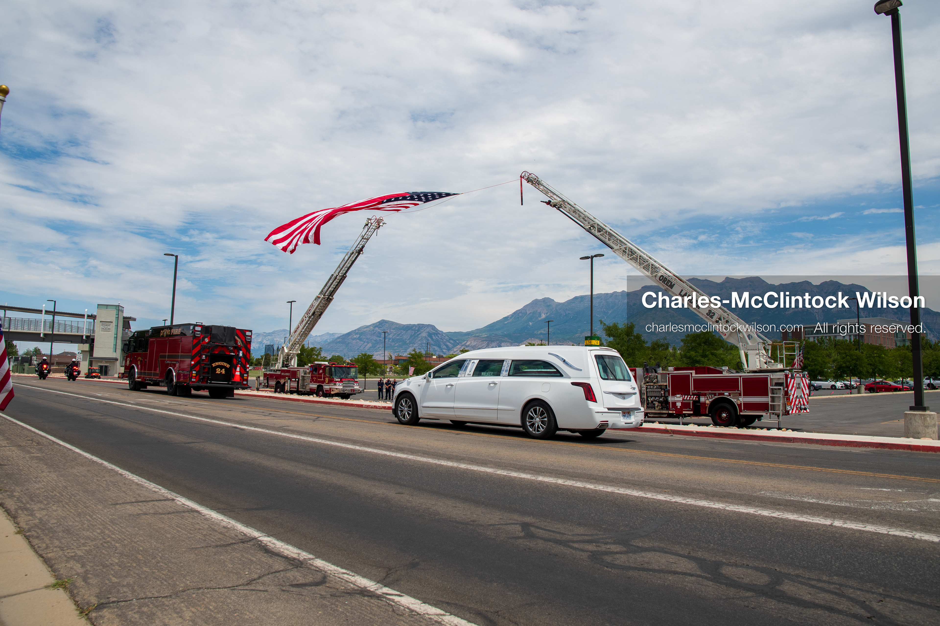 A white Cadillac hearse drives past a massive American flag suspended between two extended fire department ladder trucks during a funeral procession for Provo Fire Captain Spencer Long and his family in Orem, Utah, on Wednesday, July 29, 2026. More than 300 people came to the public memorial ceremony to honor the five family members who died after a flash flood hit their campsite near the Sunglow Campground in Wayne County. (Photo by Charles-McClintock Wilson/Sipa USA)