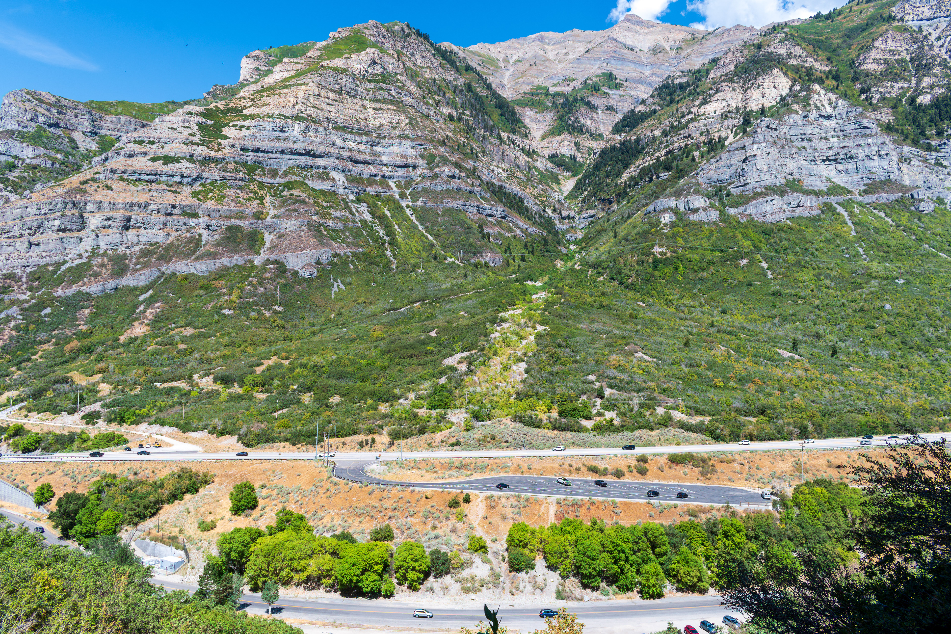 Provo, Utah, US — Sept 1, 2025: A scenic view from Bridal Veil Falls captures steep, layered rock formations and lush vegetation, with vehicles winding through the rugged terrain along U.S. Route 189.