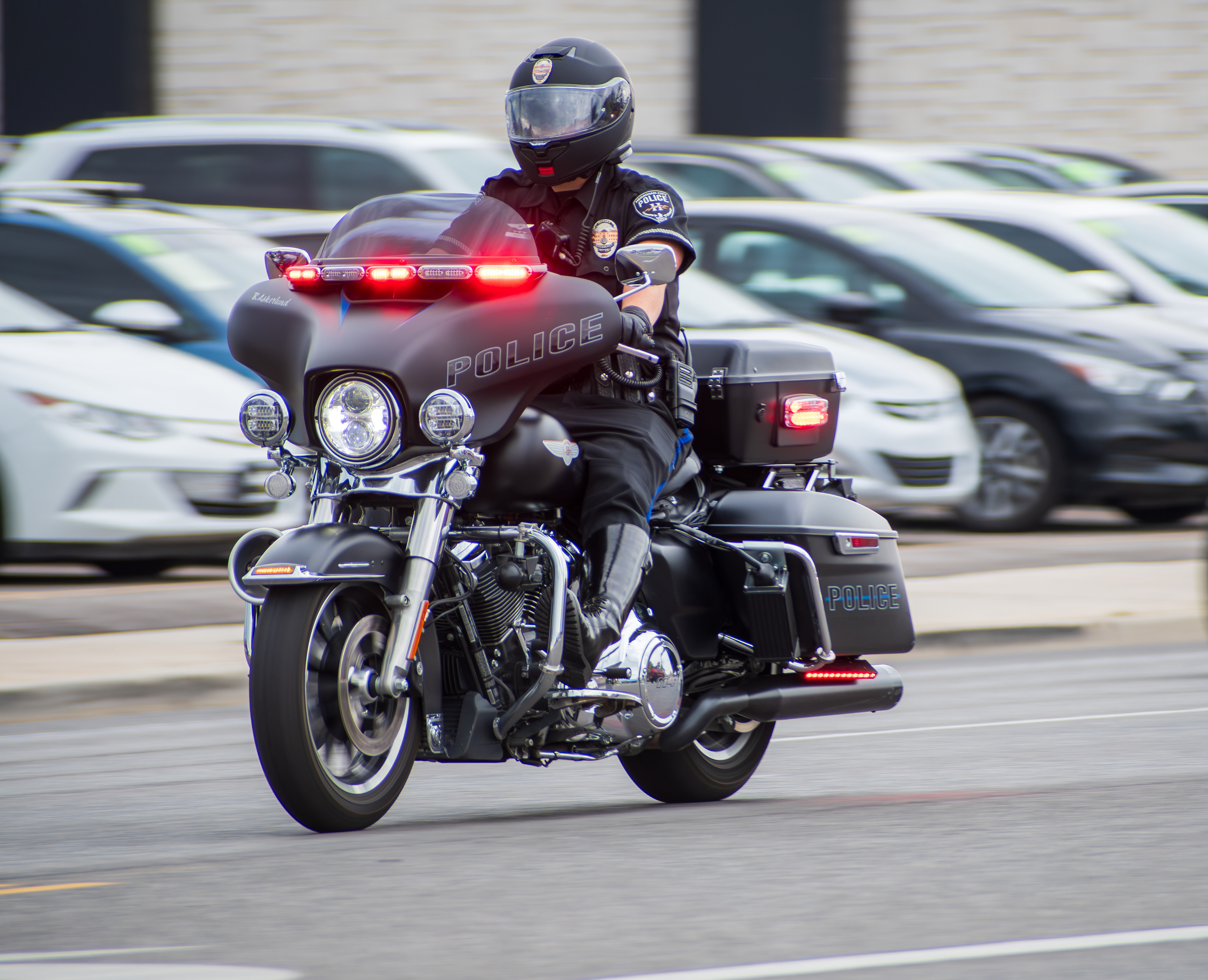 Murray, Utah — Sept. 8, 2025: A police officer rides through a city street on a motorcycle with emergency lights activated, navigating urban traffic near a row of parked cars.