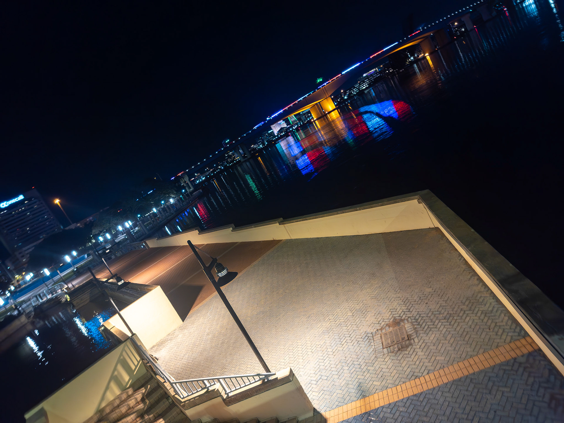 A mix of warm-toned streetlights and cooler ambient lighting creates a layered lighting effect along the riverwalk. The vibrant reflection of the Main Street Bridge’s colored LEDs in the St. Johns River adds visual contrast and energy to the calm, open space in the foreground.