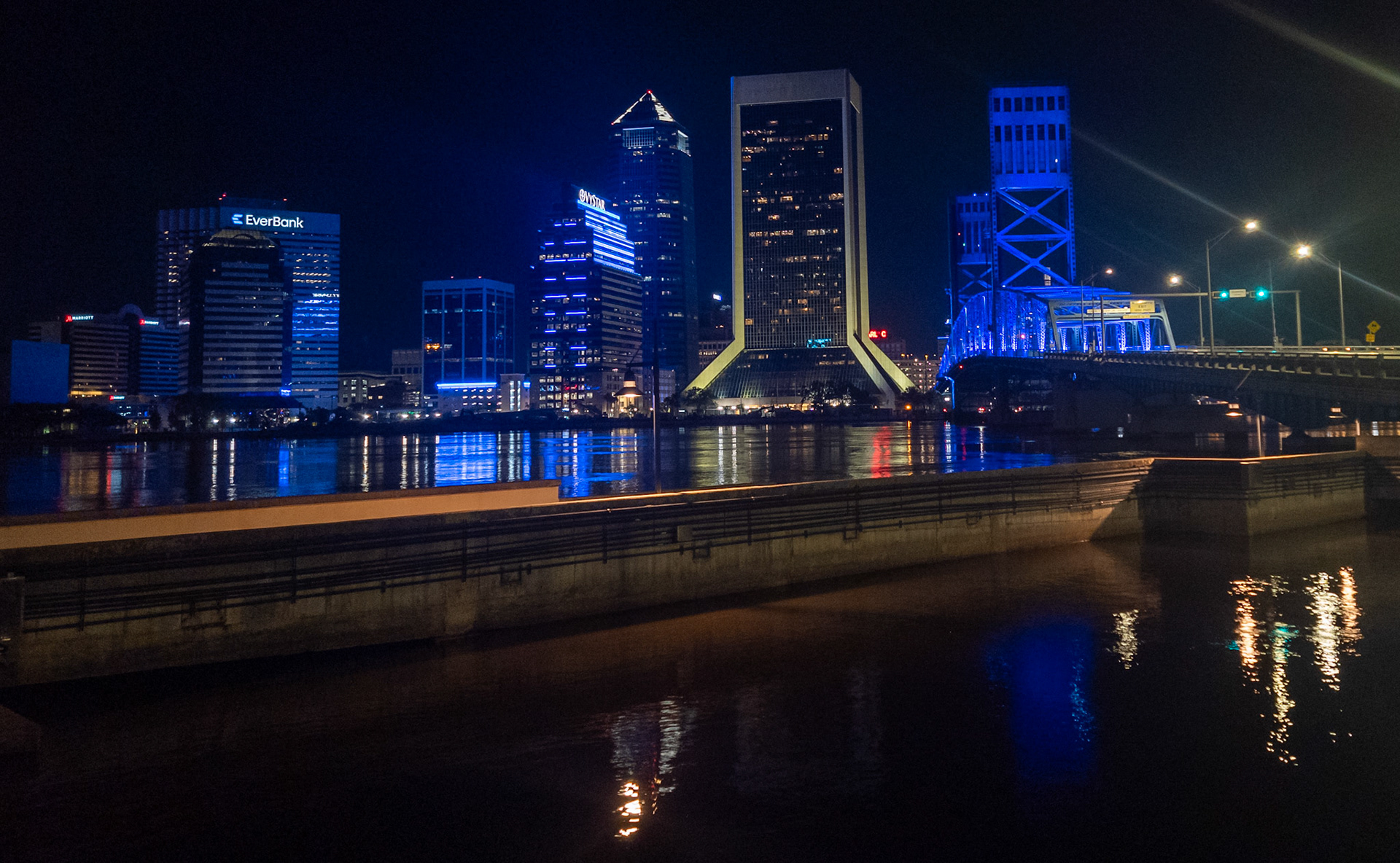 Downtown Jacksonville’s skyline glows in dramatic blues, casting crisp reflections across the river. The combination of cool LED architectural lighting and warm street lamps along the bridge produces a striking temperature contrast, amplifying the sense of depth and urban texture.