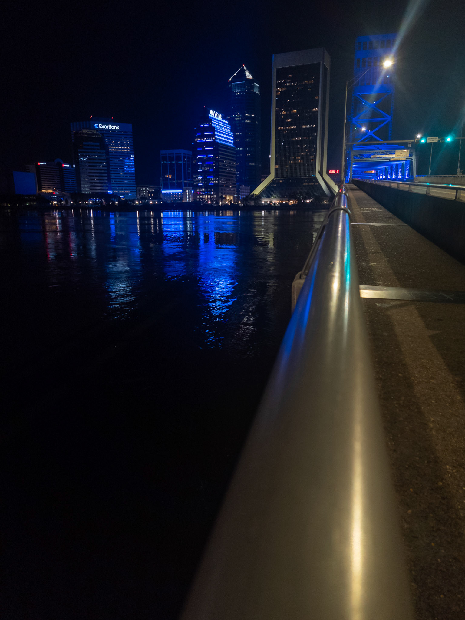Shot from a low angle at street level, this image emphasizes the stark directional lighting of streetlamps and the intense blue floodlights along the bridge. Lens flares and reflections on the rail add visual rhythm, showcasing how hard light sources can sculpt form and focus.