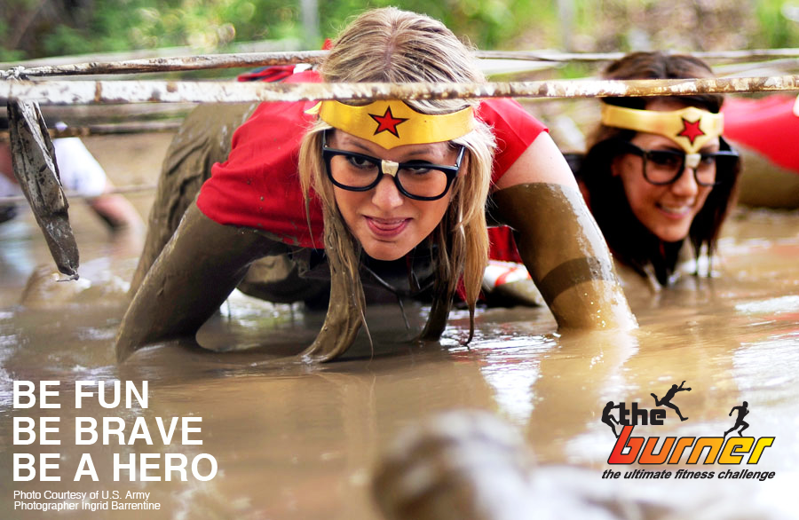 Two women crawl through mud obstacle wearing Wonder Women costumes with the text "Be Fun. Be Brave. Be a Hero" for The Burner