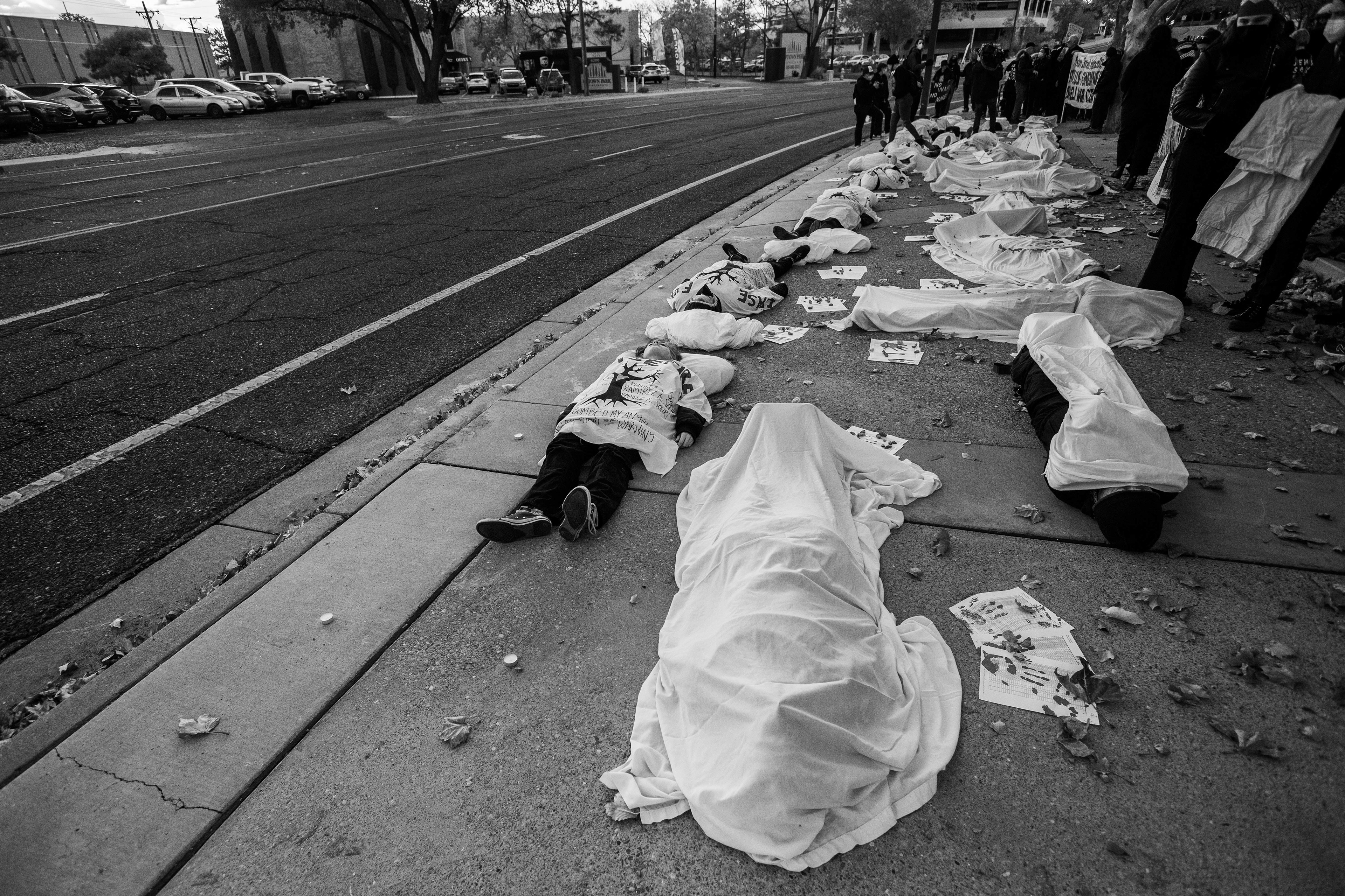 A stark line of participants lies on the ground, covered in white sheets marked with red, outside Representative Melanie Stansbury's office, as a grim reminder of the lives lost in the recent Israel-Palestine conflict.