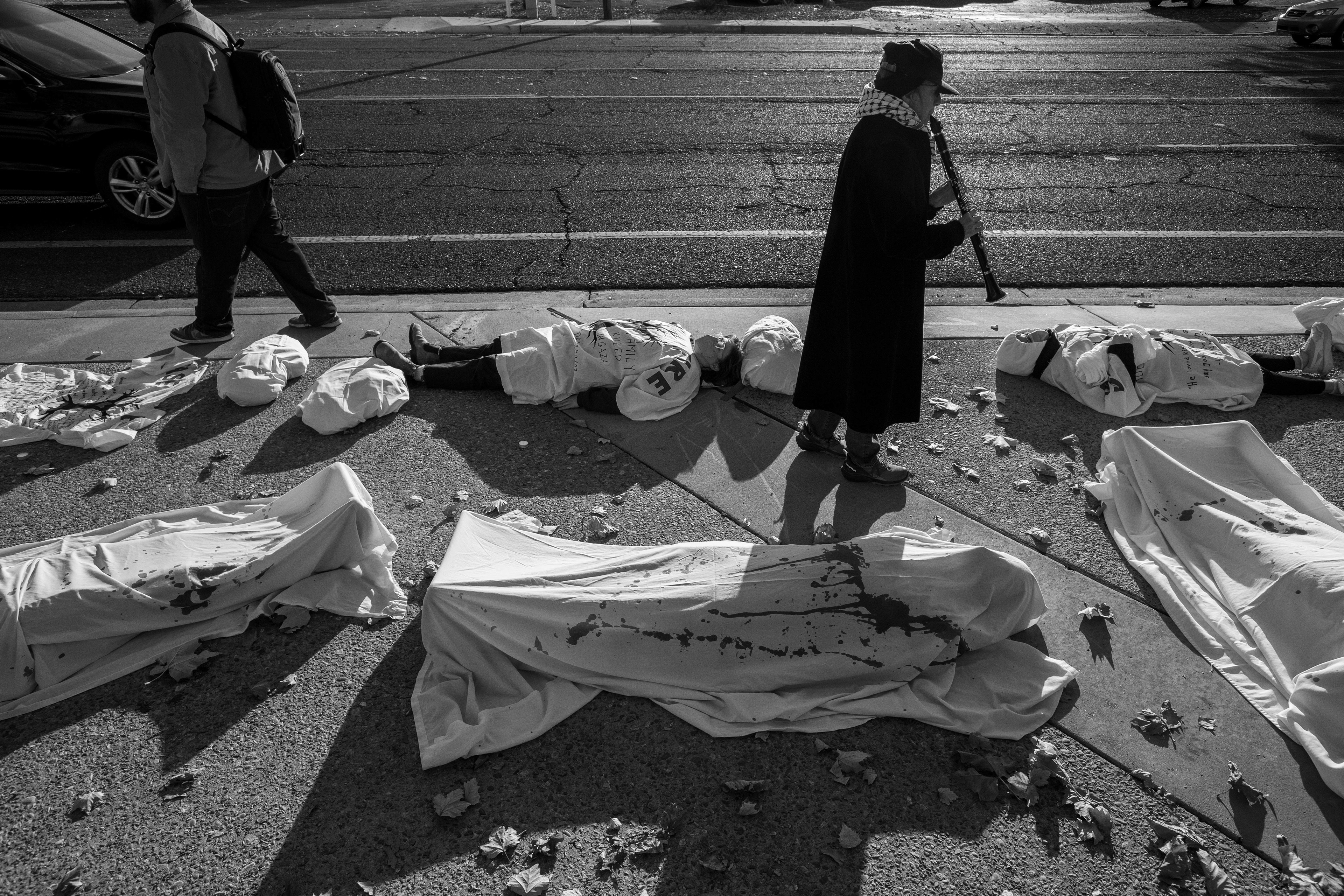 A stark line of participants lies on the ground, covered in white sheets marked with red, outside Representative Melanie Stansbury's office, as a grim reminder of the lives lost in the recent Israel-Palestine conflict.