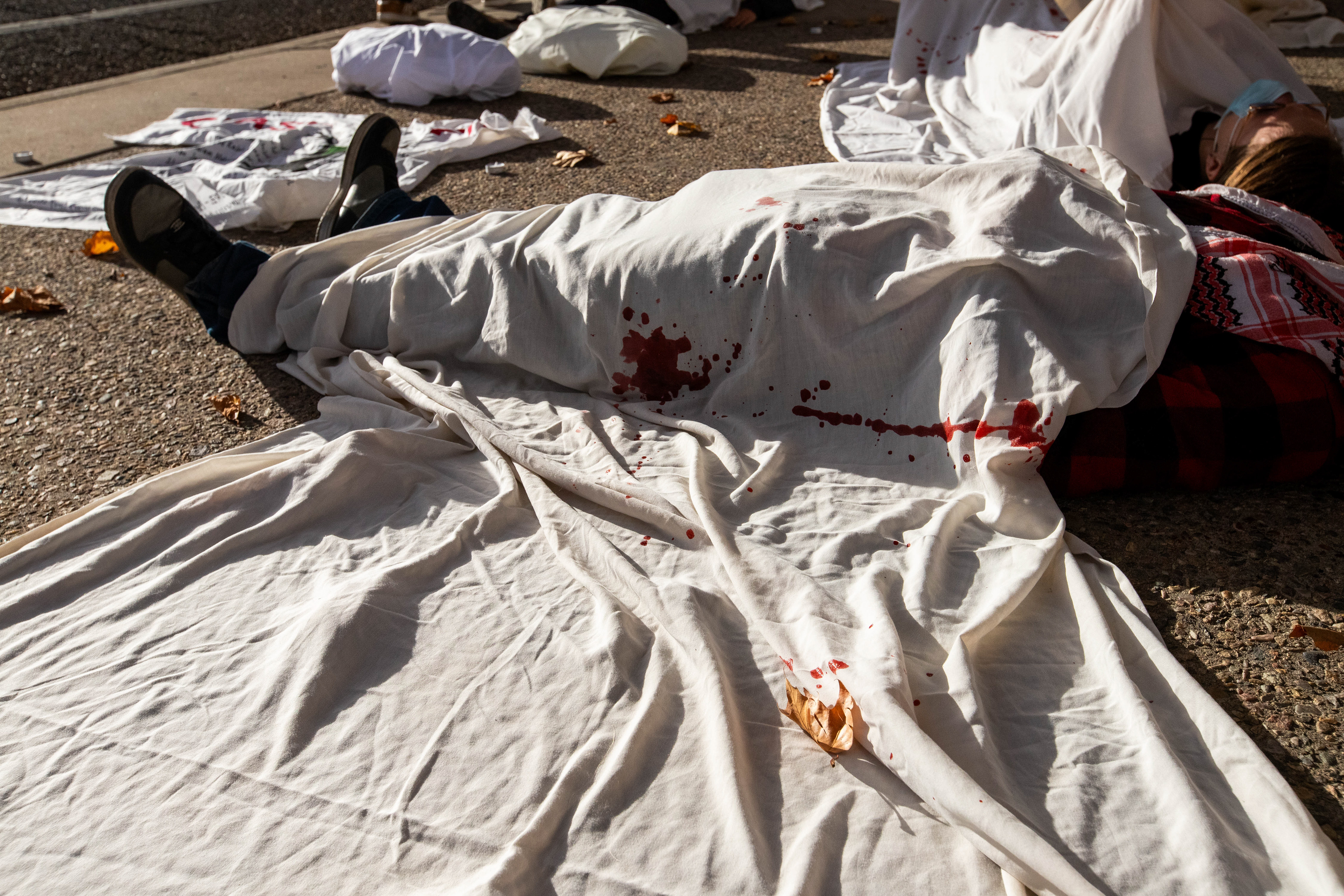 A participant lies on the ground, covered in white sheets marked with red, outside Representative Melanie Stansbury's office, as a grim reminder of the lives lost in the recent Israel-Palestine conflict.
