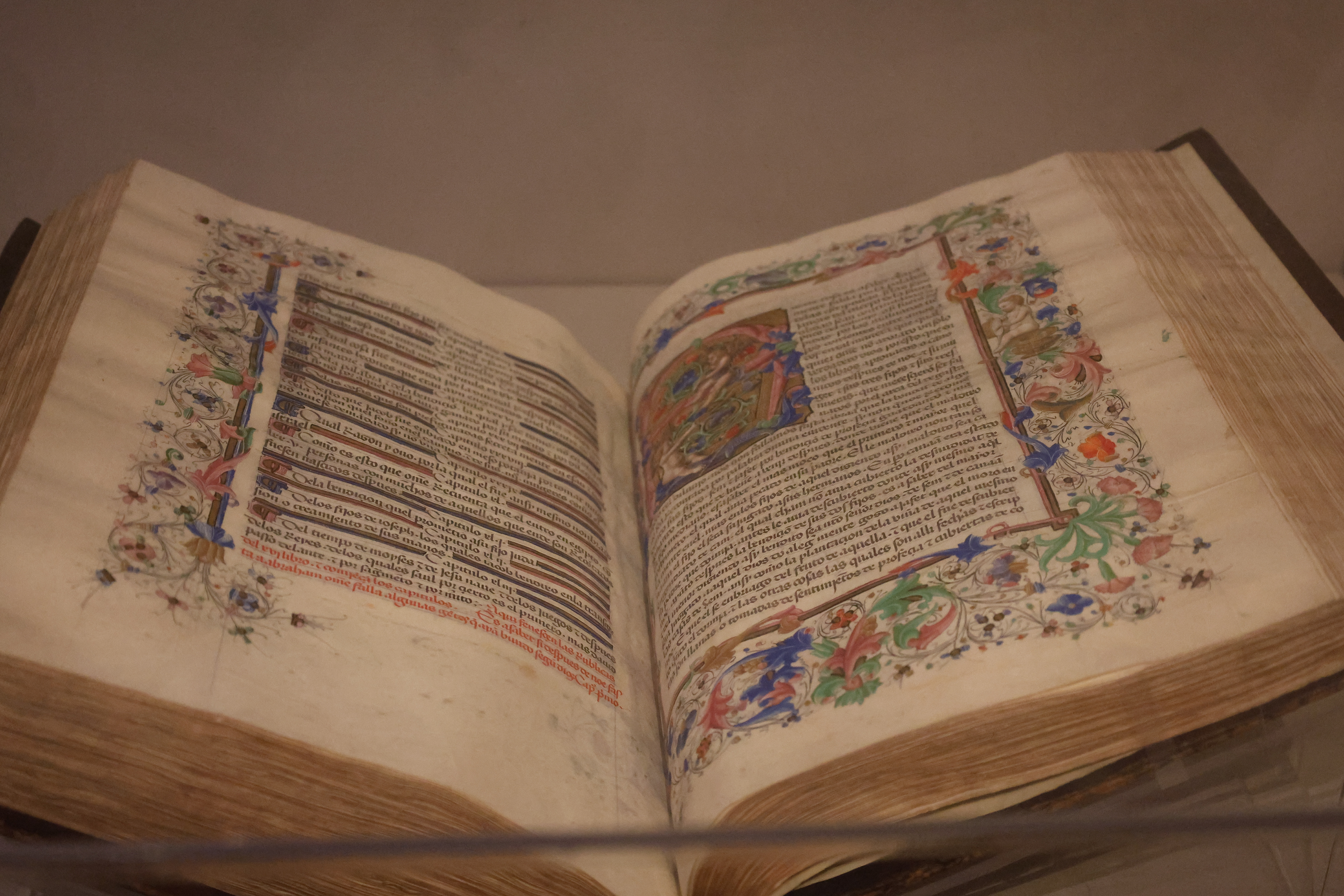 An illuminated manuscript open to a spread with elaborate floral borders in blue, green, red and gold, dense handwritten text in black and red ink, and a painted miniature at the upper right of the right-hand page.