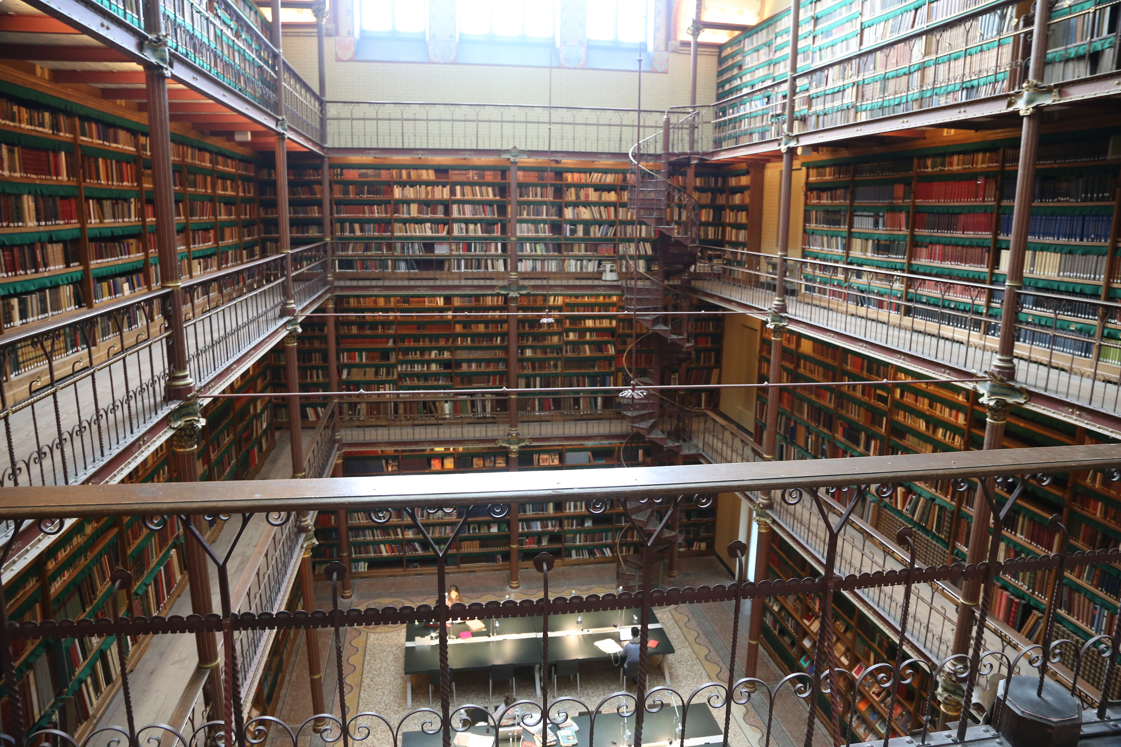 The Rijksmuseum Research Library in Amsterdam, seen from an upper gallery level — four floors of floor-to-ceiling bookshelves on all sides, dark ironwork balustrades, ornate spiral staircases, and two researchers working at a lit table on the ground floor far below.