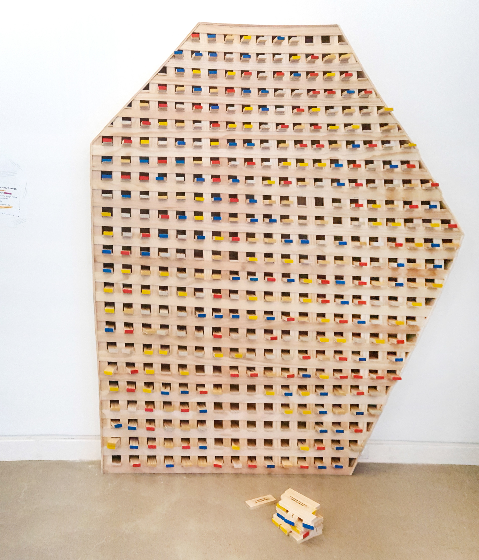 A light wood sculpture stands in front of a white wall with white trim at the bottom on a grey cement floor. The sculpture is shaped as an irregular hexagon and it is inspired by the shape of the monument Rosetta Stone. The facade of the sculpture is hollow and covered by a grid of light wood strips leaving holes or shelves in the facade. In the holes are pegs with edges painted in yellow, blue and red.