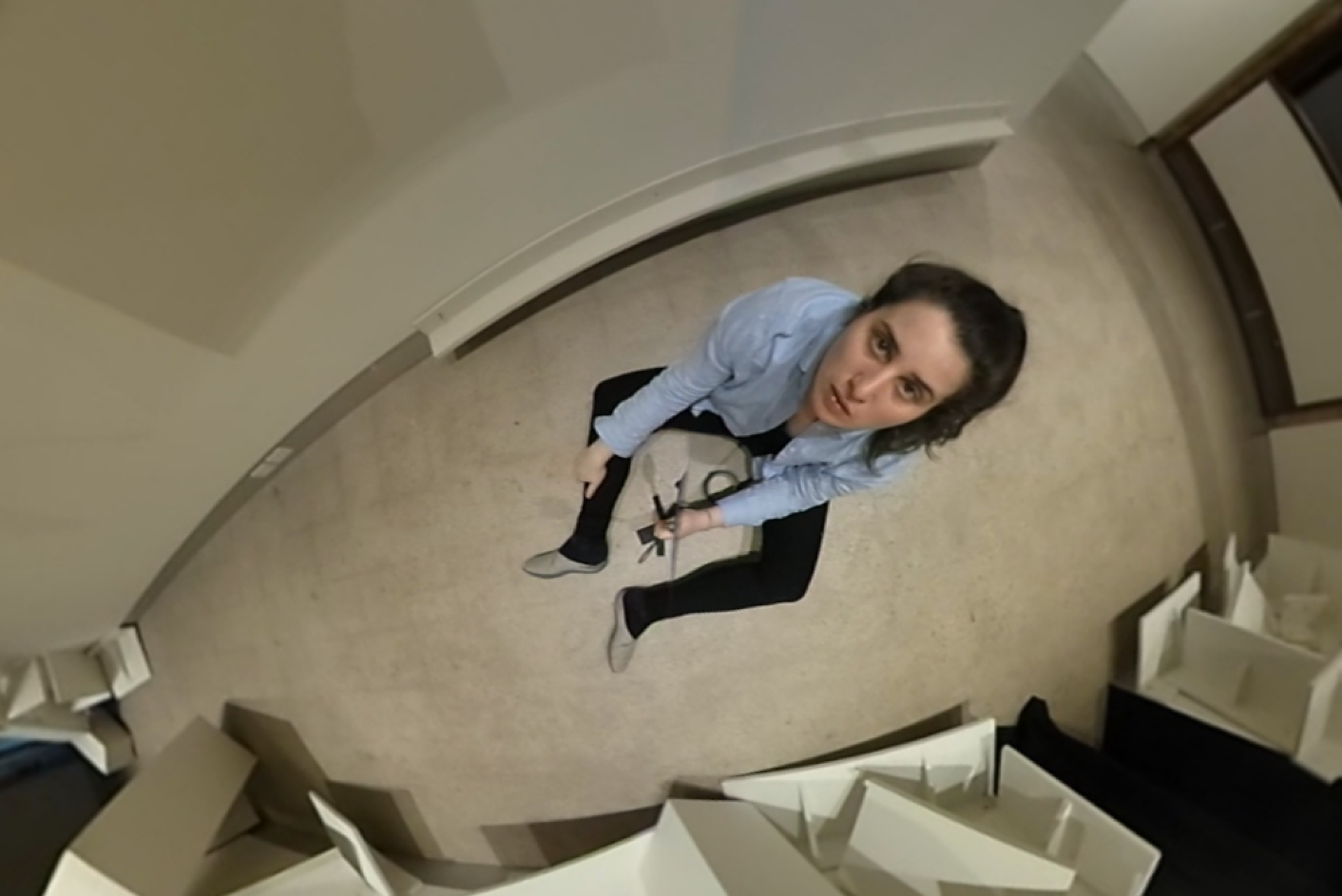 Zoe sits on a grey cement floor. They are wearing a blue long sleeve shirt, black long leggings, and grey slippers. They have long dark hair in a messy pony tail. The photo is taken with a fish eye camera positioned over Zoe. Zoe is looking into the camera with a surprised face.
