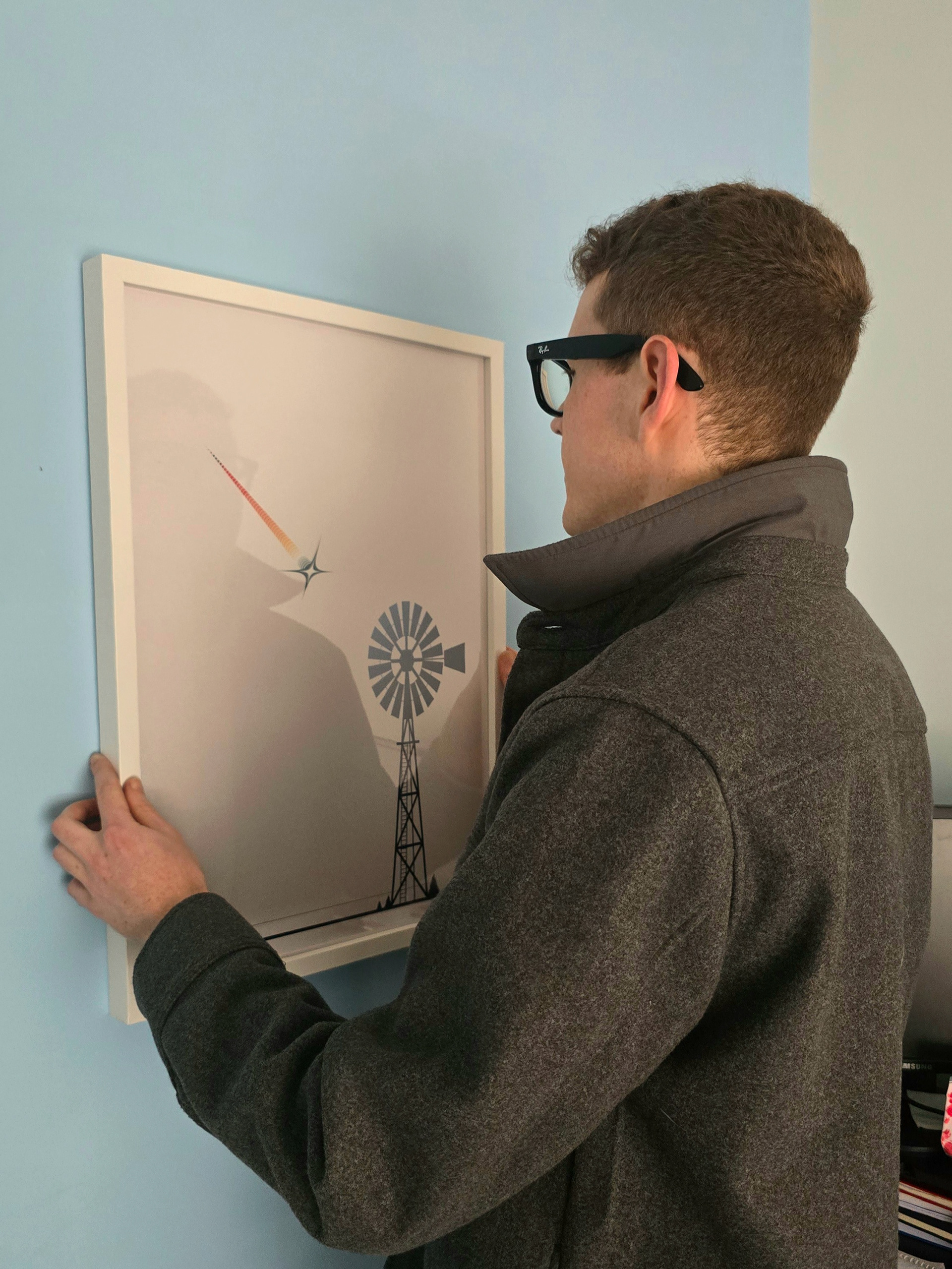 The artist, LX, hanging a white-framed print of the Stillness (Before the Spark) design from the Ascent to the Stars collection on a blue wall.
