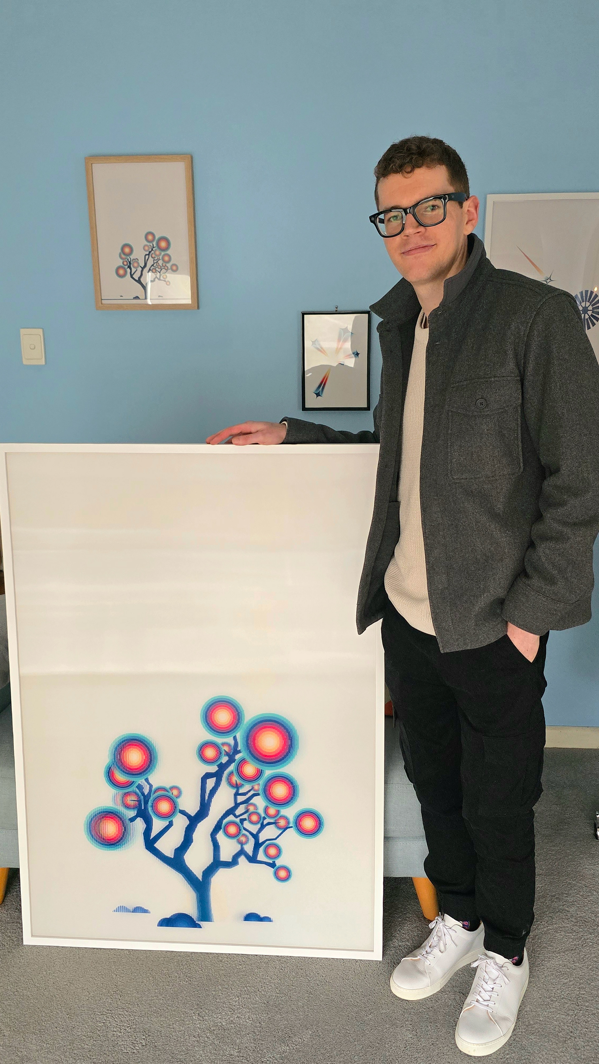 LX, the artist, standing next to a large, framed lenticular print of the Hakea Tree from the Ascent to the Stars collection, emphasising the print size.