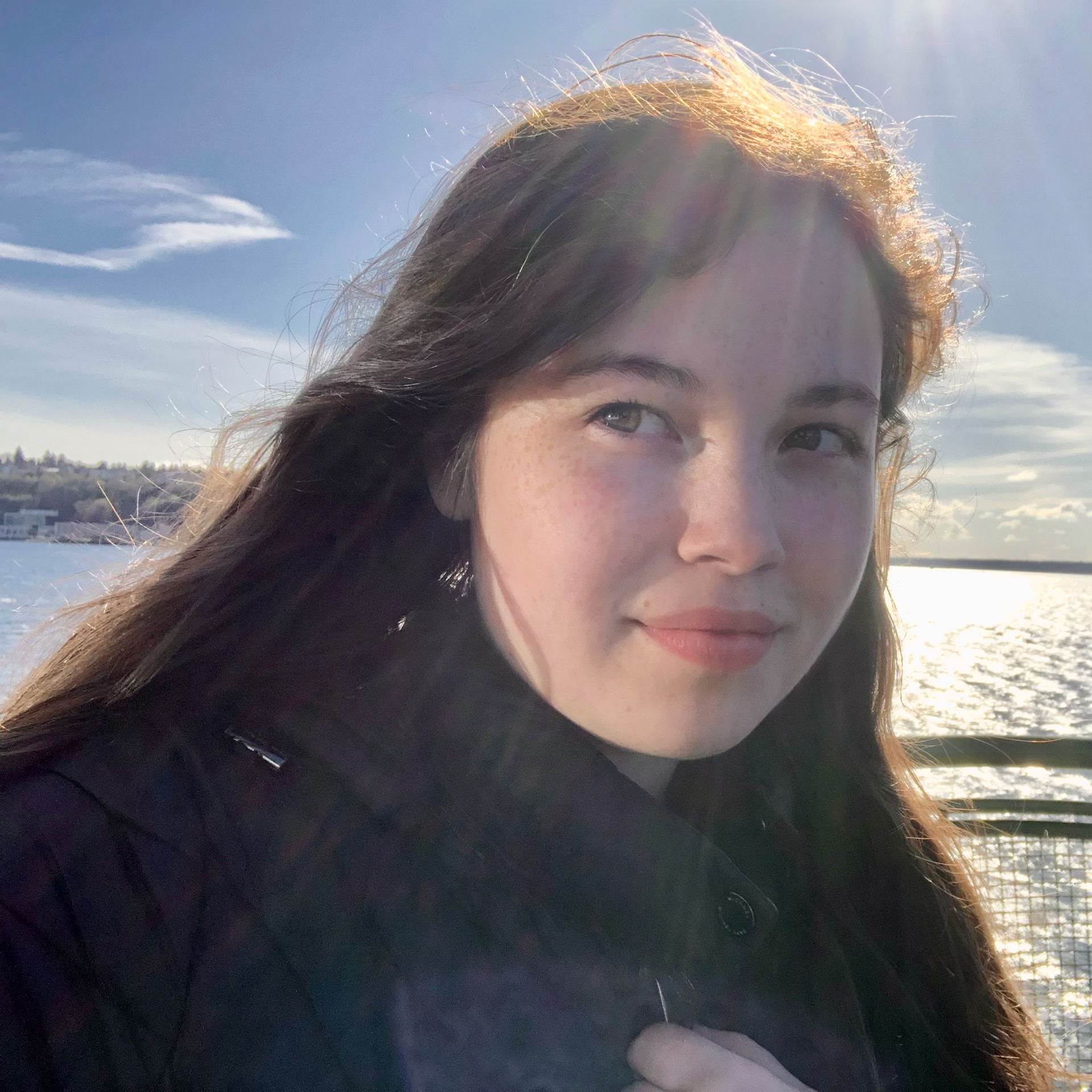 Portrait of young white women with brown long hair and freckles. She is if front of a big body of water and the sun light is shining on her ( creating glare with the camera). She is looking past the camera and she has a soft grin to her face.