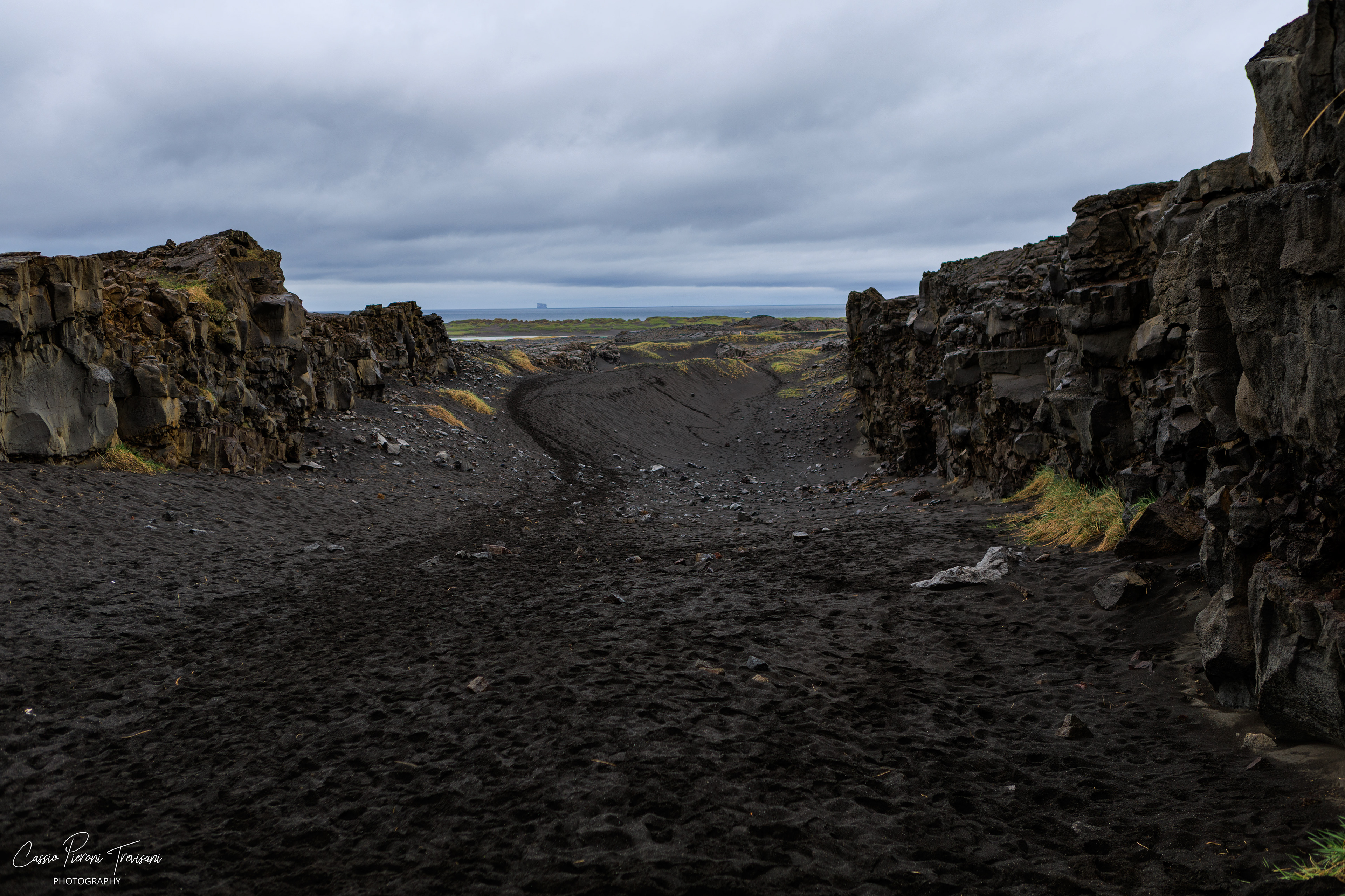 A black sand path cuts through dark lava walls near the Bridge Between Continents, leading the eye toward the Atlantic horizon.