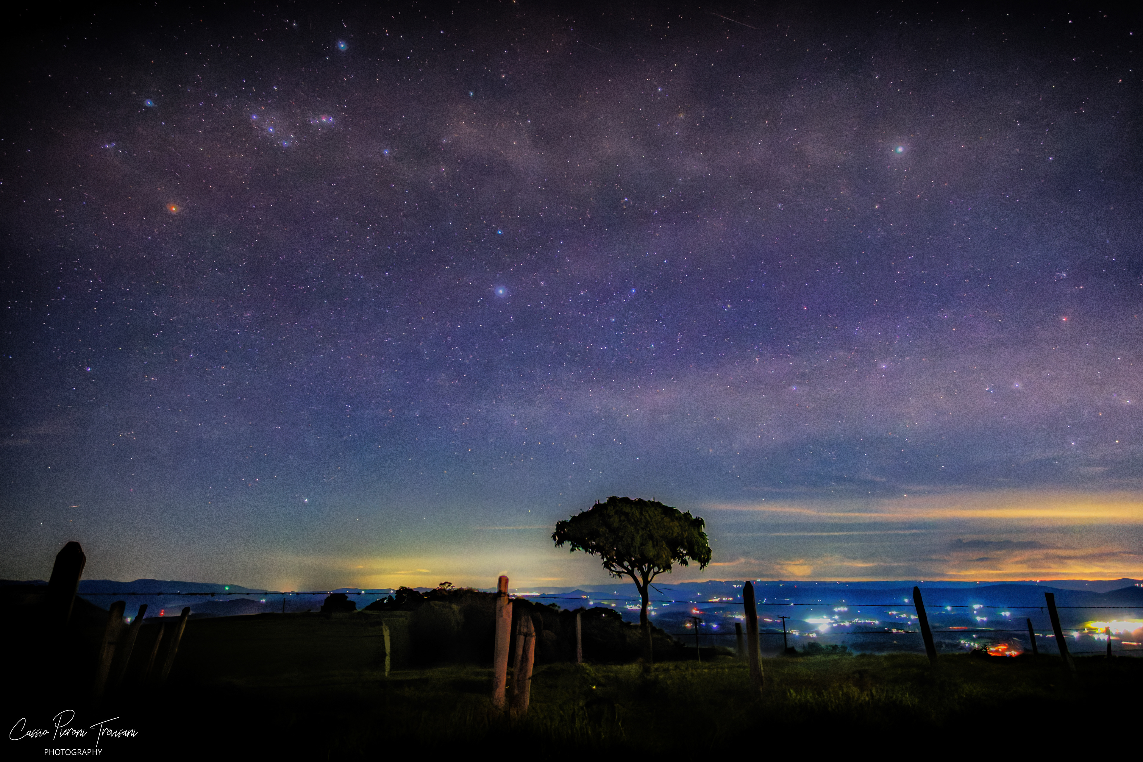 Milky Way stretching across the night sky above a lone tree and rural landscape, with city lights in the distance in Jacutinga, Minas Gerais, Brazil.