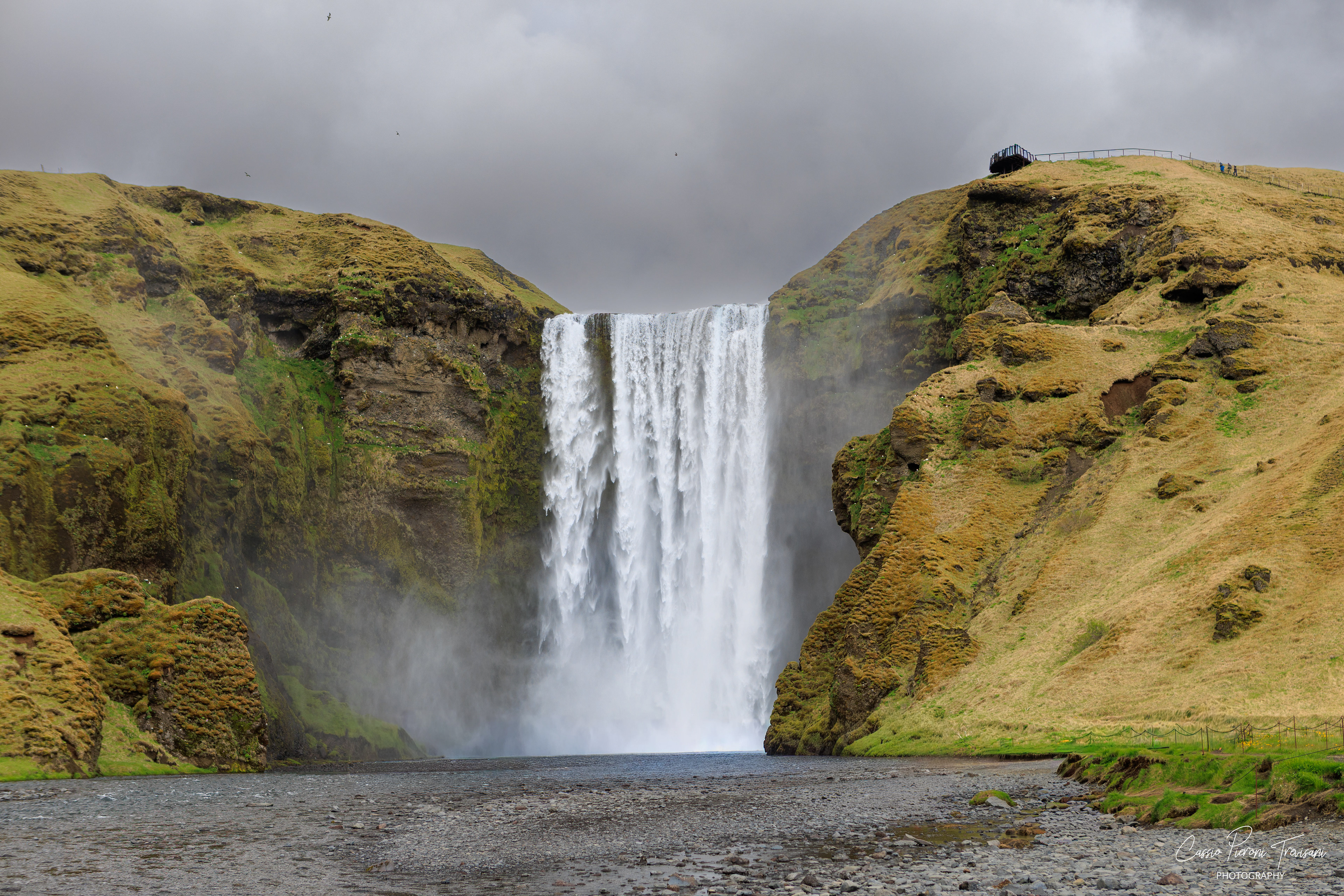 Skógafoss thunders between green cliffs, sending mist across the river stones and into the valley.