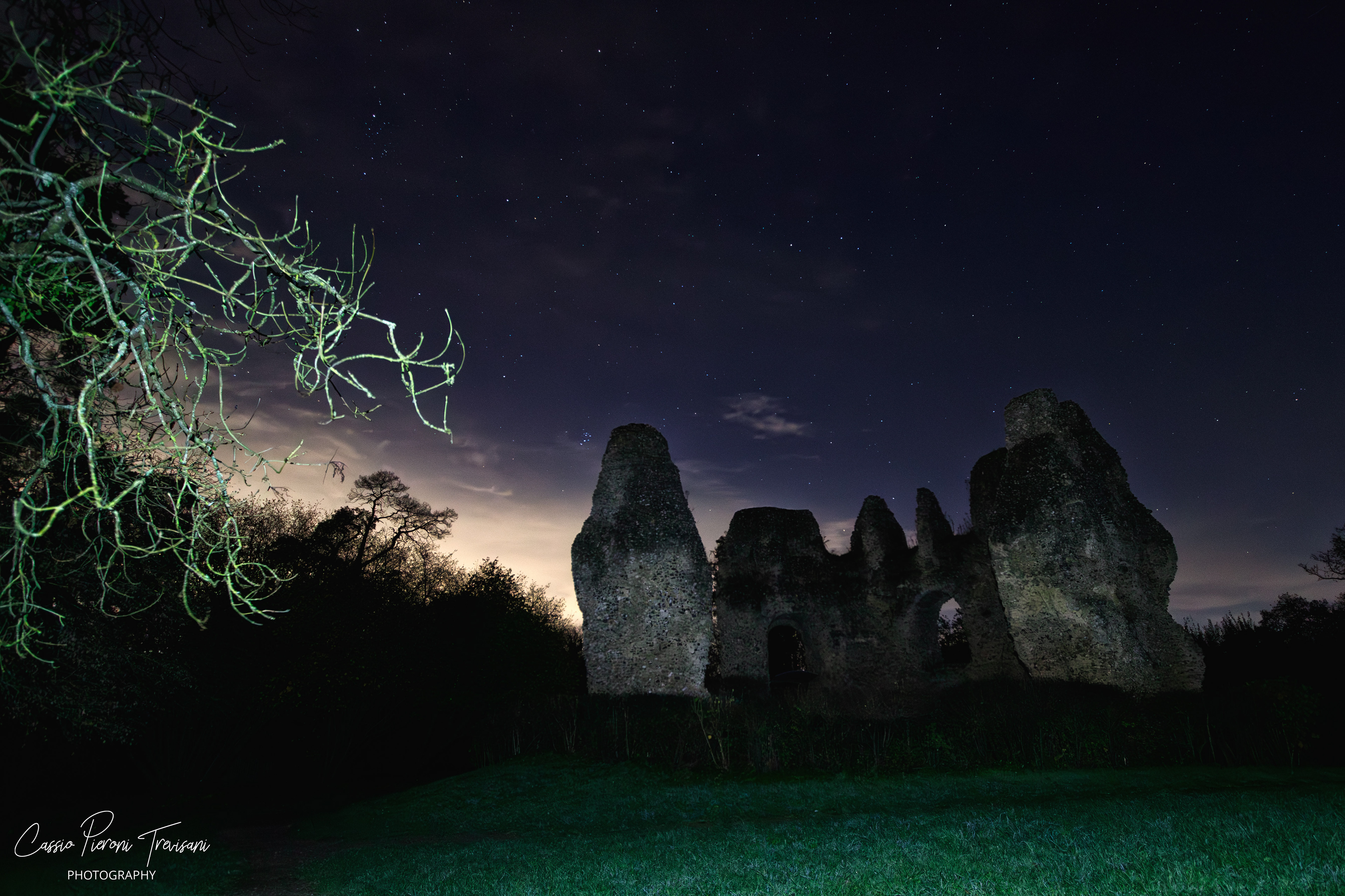 Distant view of Odiham Castle ruins at night with foreground tree branches lit against a starry sky.