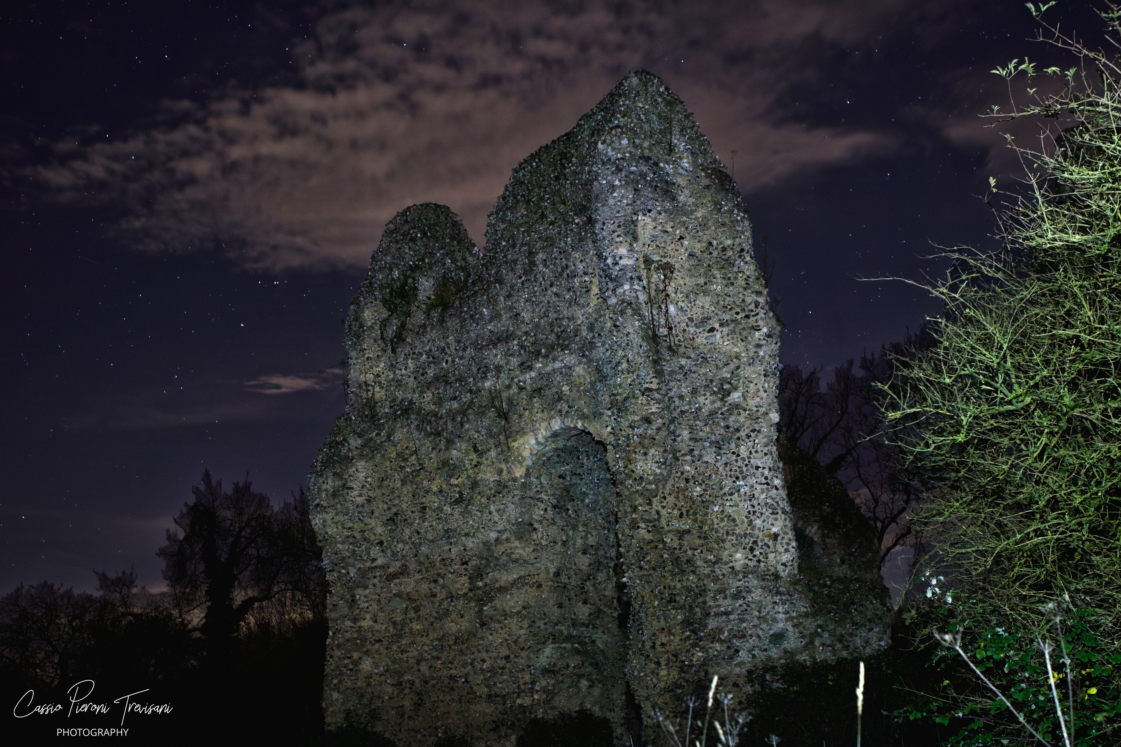 Tall stone ruin of Odiham Castle lit at night with trees framing the scene under a starry sky.