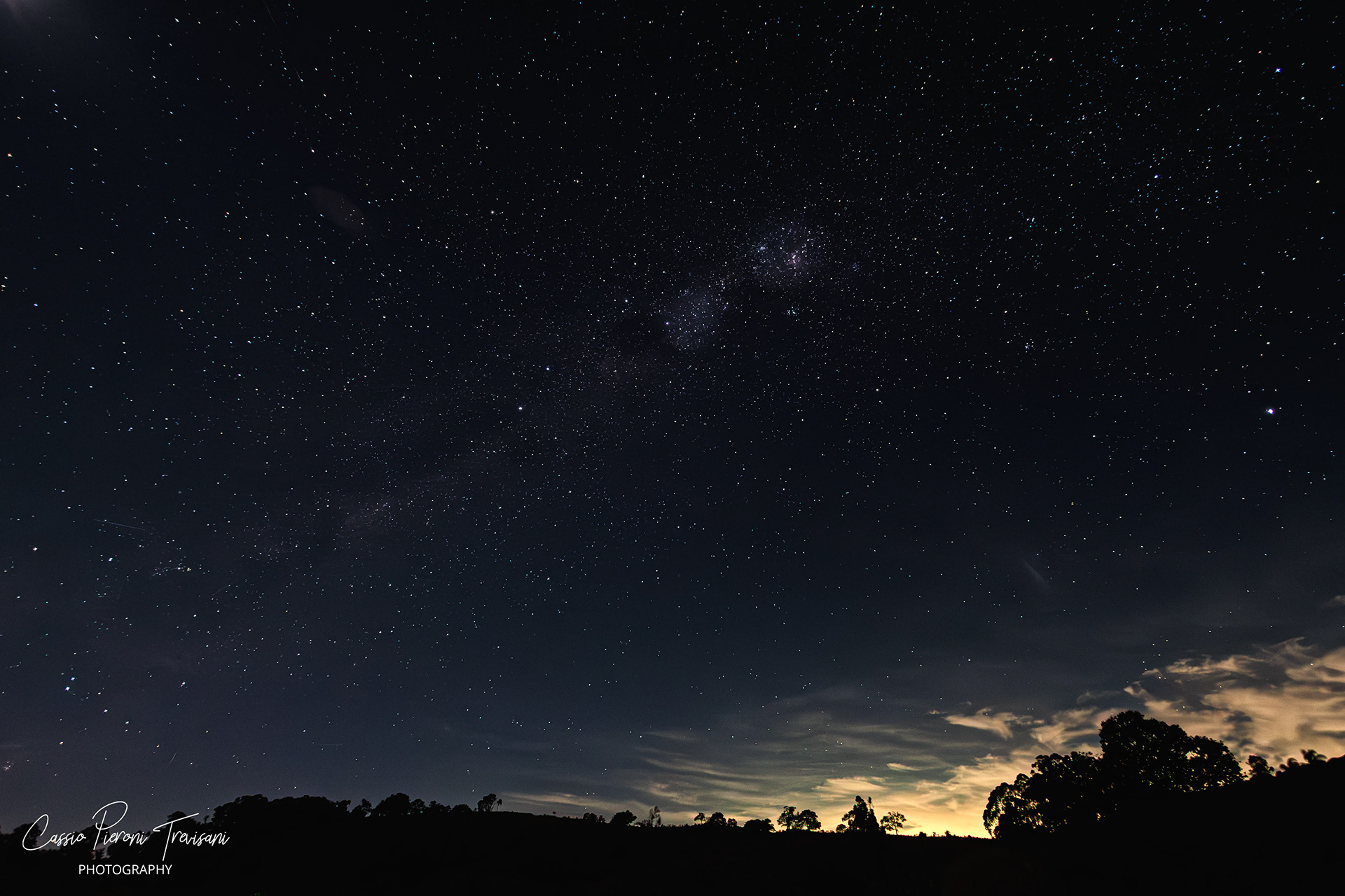 The Milky Way forms a subtle diagonal band across a dark, cloud-dusted night sky above silhouetted hills in rural Minas Gerais.