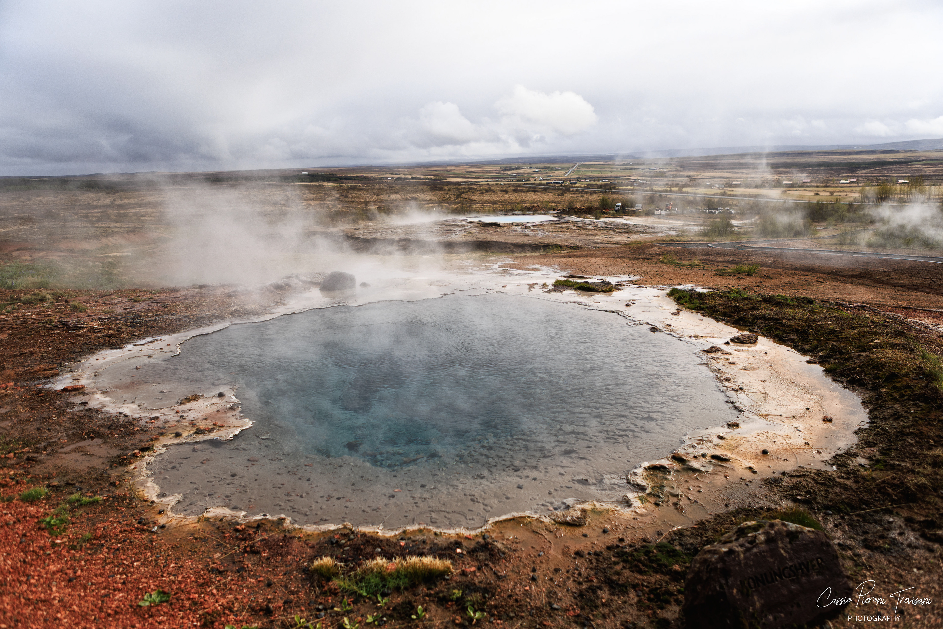Steam rises from Konungshver, a vivid geothermal pool surrounded by mineral earth and the open landscape of Haukadalur.