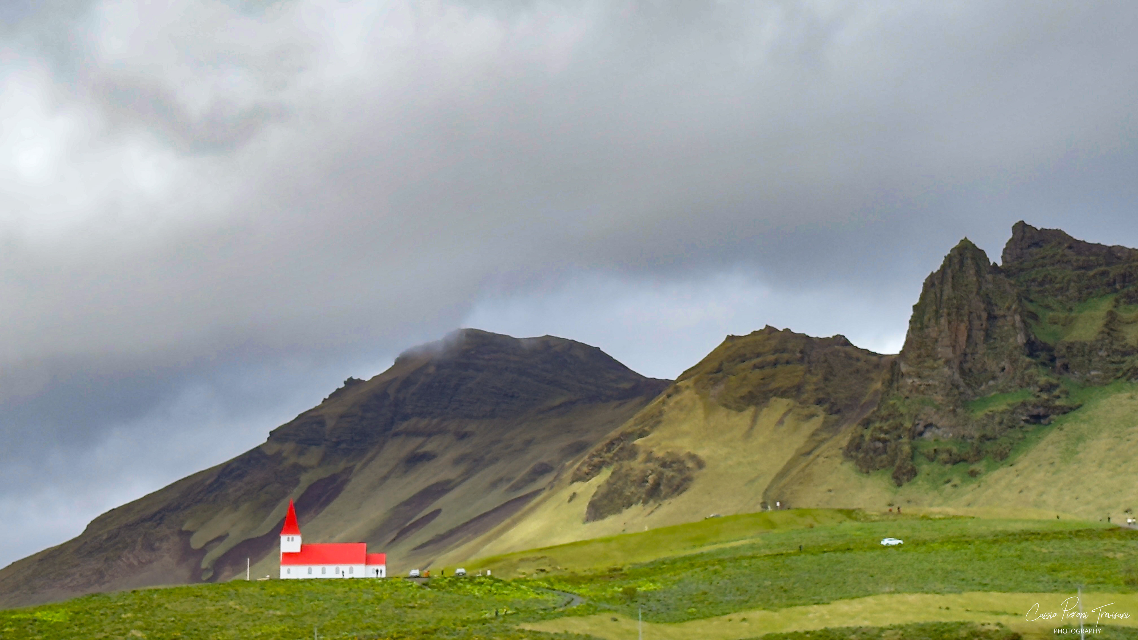 Víkurkirkja rises above the village of Vík, its red roof standing bright against the green slopes and dark volcanic cliffs of Iceland’s South Coast.