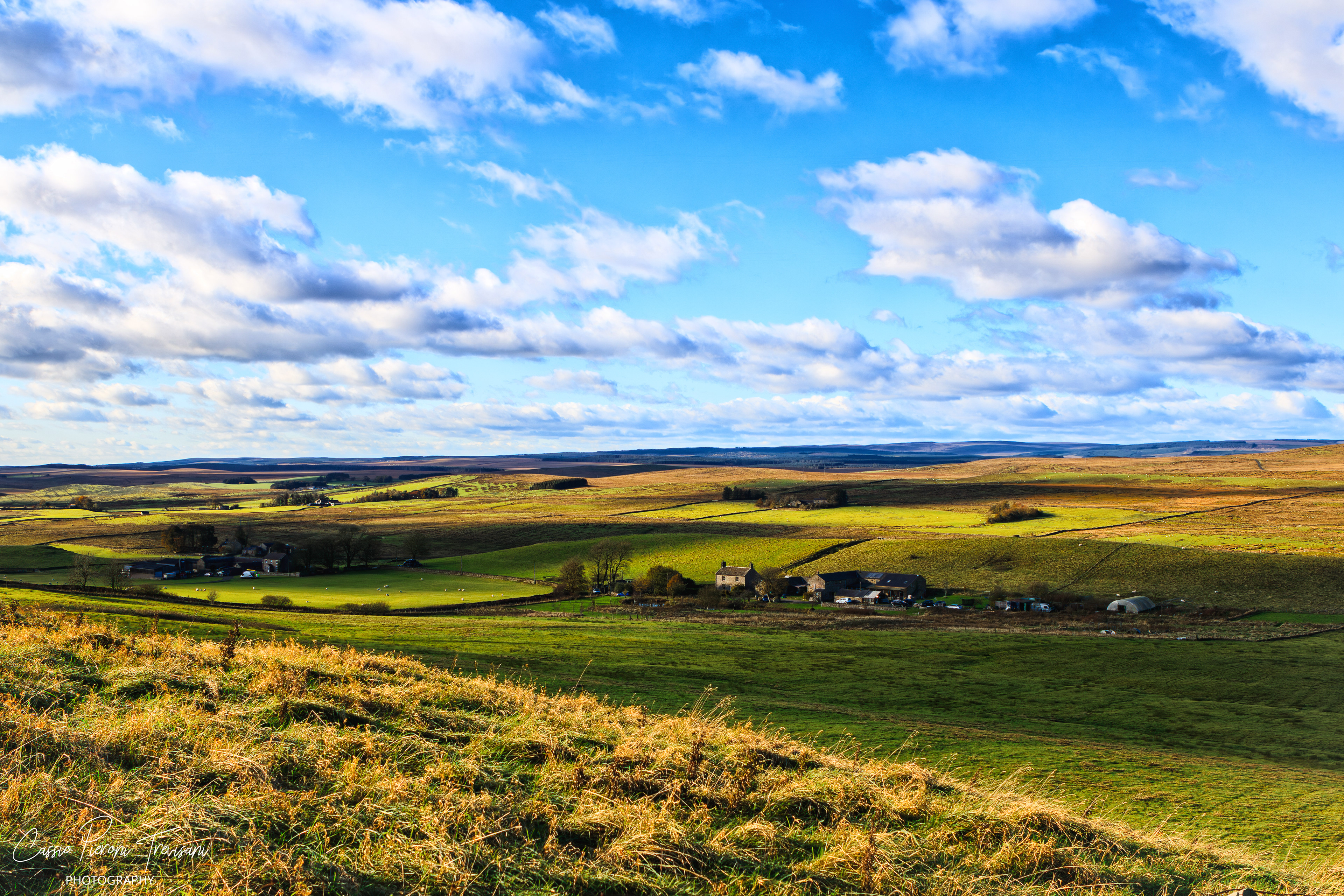 Golden autumn grass in the foreground gives way to rolling farmland and distant hills under a wide, cloud-filled sky.
